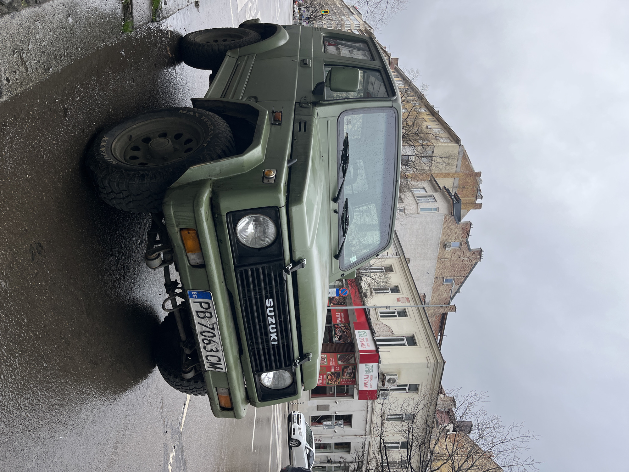 The image shows a green Suzuki jeep parked on a wet city street. The jeep has a visible license plate reading "PB 7063 CM", indicating it's registered in Bulgaria. The background features typical European architecture, including a building with a red and white awning and signage in Cyrillic script advertising "Гурман". The scene suggests a cloudy day in Sofia, Bulgaria. A white sedan is partially visible behind the jeep.