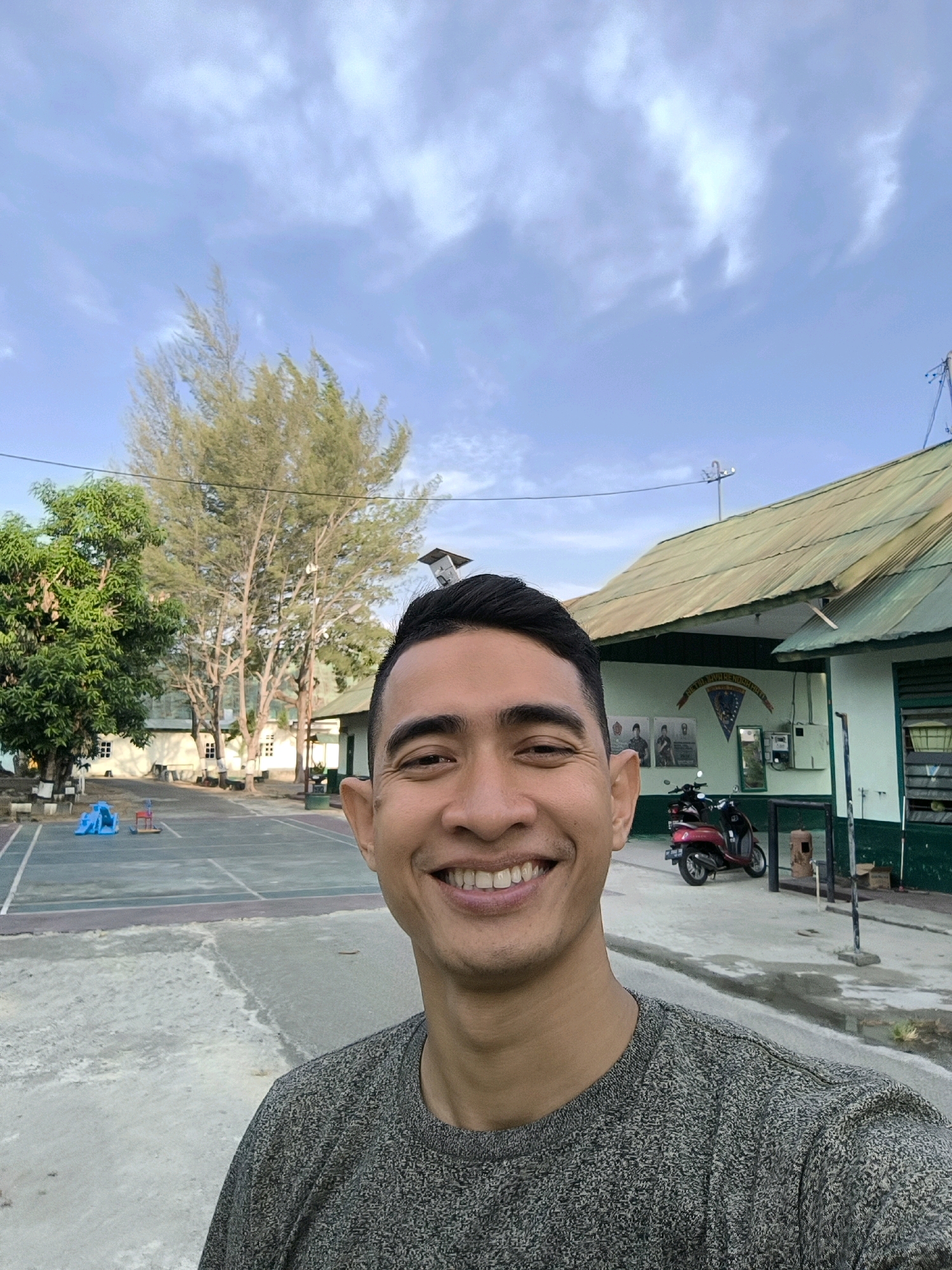 A man with short, dark hair and a wide smile takes a selfie outdoors. He is wearing a grey, textured t-shirt. Behind him, a paved courtyard is visible, with a basketball court marked out. Several trees of varying sizes are scattered around, including a large, mature pine tree. In the background, a long, light green building with a corrugated metal roof is present. Two scooters, one red and one dark-colored, are parked in front of the building. The sky above is a pale blue with scattered, wispy clouds. The setting appears to be a school or communal area in Gorontalo, Indonesia, judging by the architecture and vegetation typical of the region.