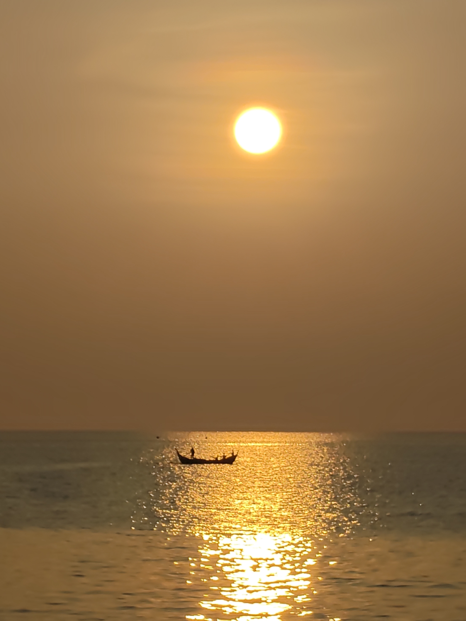 The image presents a serene seascape at either sunrise or sunset, bathed in a warm, golden glow. The sky and water are permeated by intense amber and orange hues, creating a tranquil and picturesque scene.
In the upper center of the frame, the sun is a brilliant, circular white orb, casting its radiant light across the entire vista. The sky around the sun transitions from bright yellow and orange closest to the light source to a softer, more muted peach color towards the edges of the frame, with no visible clouds.
The calm sea occupies the lower half of the image. A sparkling path of sunlight stretches from the horizon directly towards the foreground, creating a shimmering, golden reflection on the water's surface. This reflection is brightest and most defined closest to the viewer, where individual light glints are visible.
A small, traditional boat, likely a fishing vessel, is silhouetted against the bright reflection on the water, positioned in the middle distance. Several figures, also appearing as dark silhouettes, are visible within the boat, indicating the presence of people. There appear to be at least three to four individuals onboard, suggesting activity such as fishing or commuting across the waters.
The overall impression is one of peaceful coexistence between nature's beauty and human daily life, capturing a quiet moment on the water. The golden hour lighting evokes a sense of calm and beauty. This scene is depicted off the coast of Lhokseumawe, Indonesia. There is no visible text within the image.