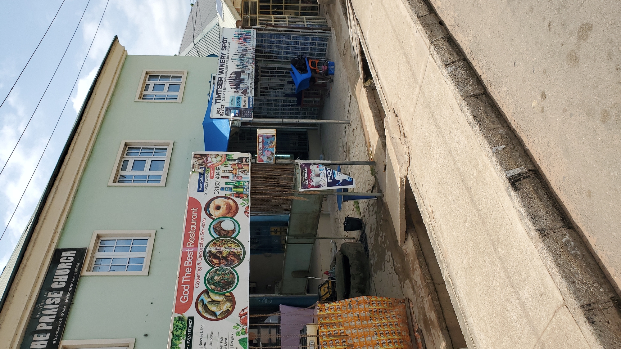 This media file depicts a street scene in Jalingo, Nigeria. The primary focus is on the exterior of a building housing various establishments. A pale green building with white-framed windows dominates the left side of the image, featuring signage for "THE PRAISE CHURCH" and "God The Best Restaurant," which advertises catering and decoration services along with a menu of food items like noodles, jollof rice, fish, meat, and doughnuts.
Adjacent to the restaurant, a business named "TIMTSER WINERY SPOT" is visible, advertising itself as a dealer and supplier of wine, beer, and soft drinks, also offering POS services, deposit, and transfer options. A blue awning shades the entrance to this establishment.
The scene appears to be set during the daytime, with natural light illuminating the area under a partly cloudy sky. The street itself is paved, with a concrete curb separating it from the sidewalk and the buildings. Several street poles are present, some displaying advertisements for "ICE CREAM" and a product labeled "POT."
Objects scattered on the sidewalk include a black bag, a deflated tire, and some discarded items. The overall impression is of a typical urban commercial street with multiple businesses operating in close proximity. There are no people prominently visible in the scene. The weather seems fair, suggesting a normal day for business. The text in the image provides specific details about the services and products offered by the businesses.