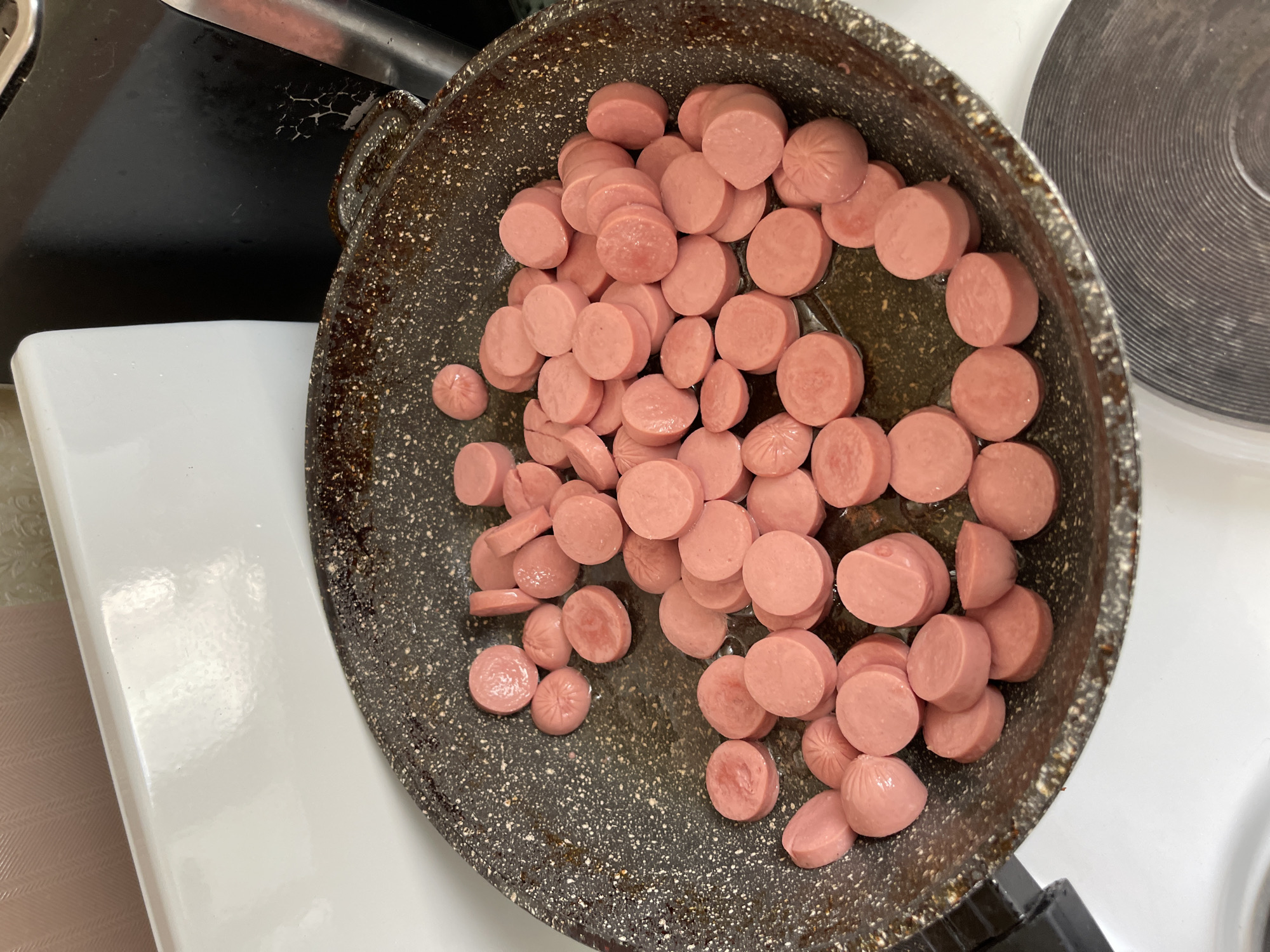 In a dimly lit kitchen, a non-stick frying pan sits on a stovetop, filled with sliced sausages. The sausages, approximately 1 cm thick, are a vibrant pink and appear to be heating in a small amount of oil or grease. The pan itself is dark grey with flecks of white and shows signs of use, with some browning and residue around the edges. The background reveals parts of a white appliance, likely a stove or refrigerator, and a textured wall. A dark, possibly stainless steel, appliance is visible to the right, partially obscuring the wall. The overall scene suggests a meal being prepared.