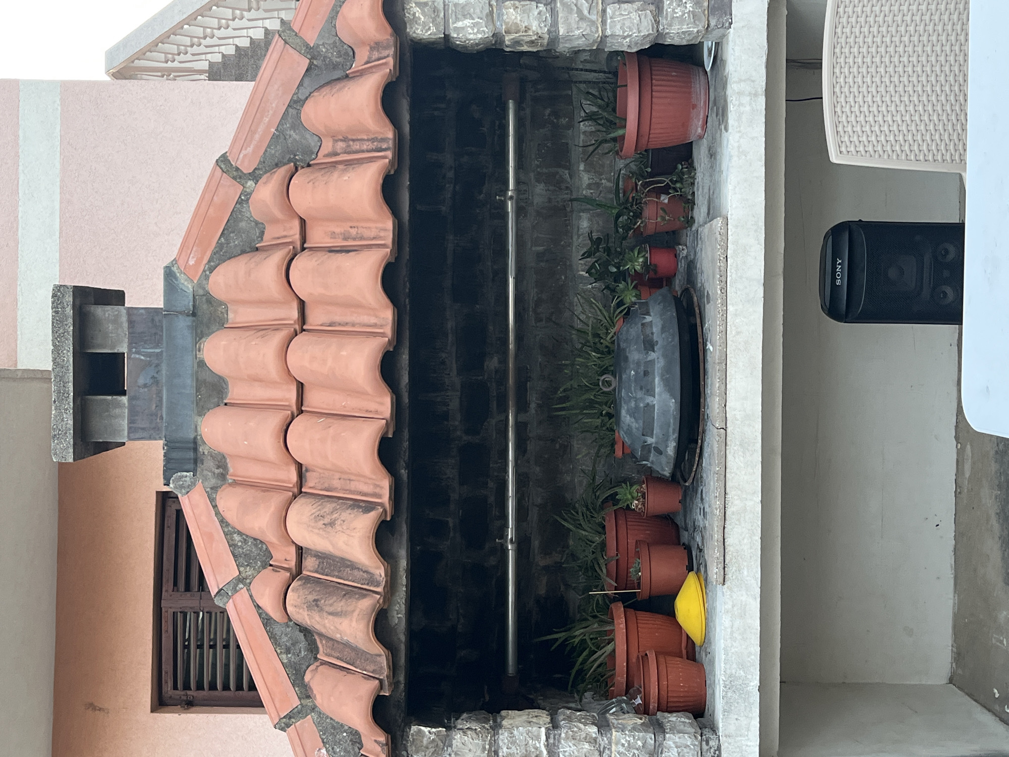 An outdoor barbecue grill with a red tiled roof is shown. The grill is made of stone and brick, with a metal rod running across the opening. Several potted plants are placed on the ledge of the grill, along with a black pot and a yellow cone-shaped object. Below the grill is a white shelf where a black Sony speaker and a chair are placed. The background shows a pink building with a window and a staircase. The scene appears to be in Budva, Montenegro.