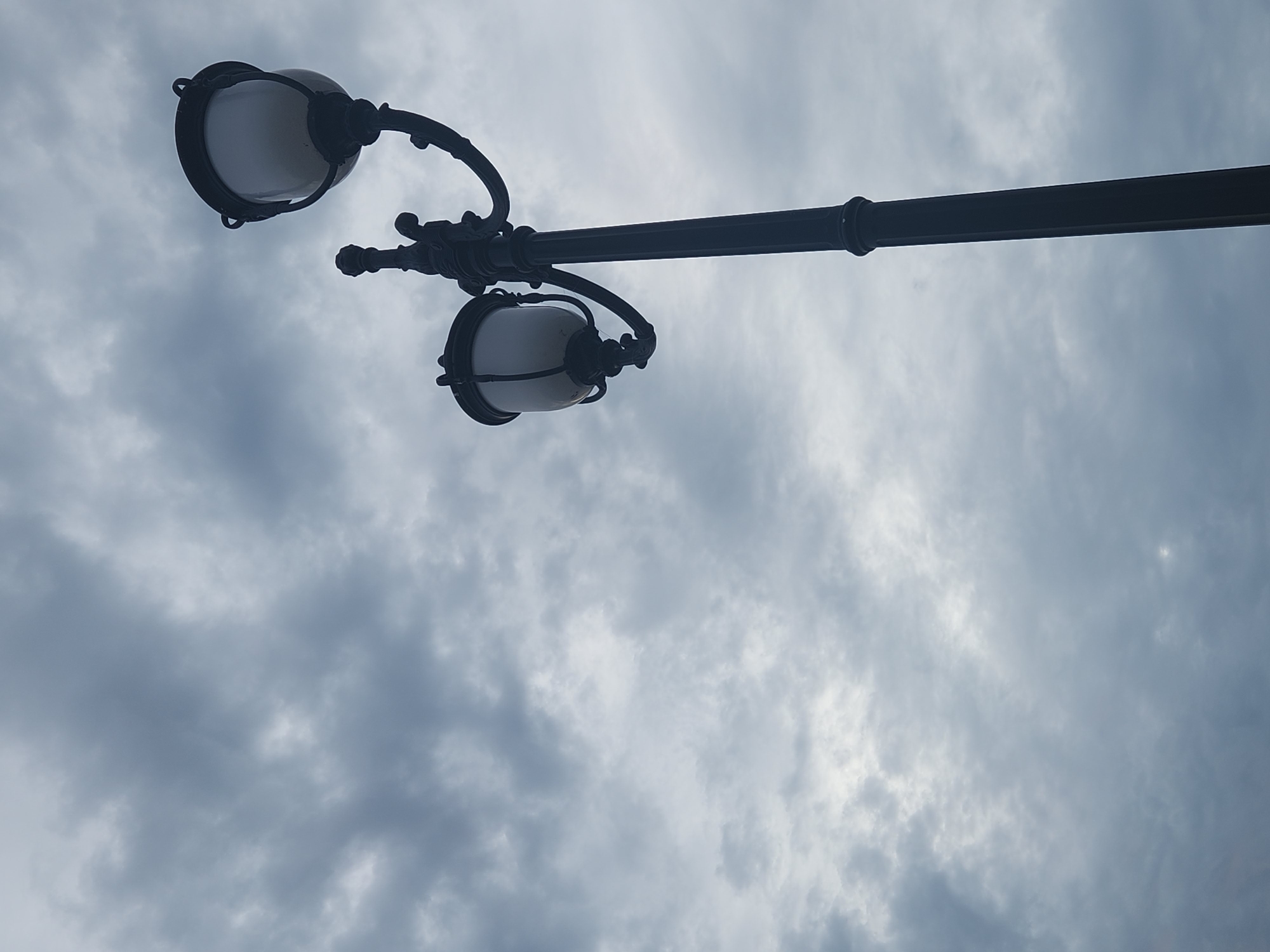 The image shows a dark, ornate lamppost with two globes, silhouetted against a cloudy, gray sky. The lamppost is positioned on the right side of the frame, with its base extending out of view at the bottom. The sky is filled with dramatic, textured clouds, giving the scene a somewhat somber or atmospheric feel. The lamppost itself appears to be made of cast iron, with intricate detailing on the arm that holds the upper globe and decorative elements along the pole. The globes are illuminated from within, though the light is soft and diffused due to the cloudy conditions. The composition is a low-angle shot, looking up at the lamppost and sky. There are no people or other objects visible in the frame. The scene does not provide any specific visual cues to place it in Altamonte Springs, United States of America.