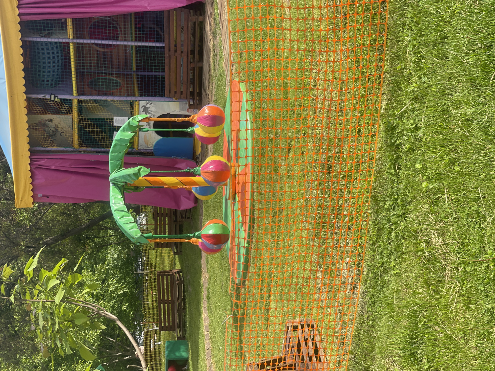 This image captures an outdoor children's play area, likely part of an amusement park or recreational facility in Poltava, Ukraine, on a bright, sunny day.
The foreground is dominated by a vibrant orange plastic safety fence, marking a boundary within a grassy field. Beyond the fence, a creative, colorful structure stands upright, resembling a stylized green palm tree. Its three "fronds" each support two large, spherical objects, colored in bright stripes of pink, yellow, blue, orange, and green, evoking a whimsical, lollipop-like appearance.
In the midground, lush green grass extends, with some scattered wooden benches or tables visible. The background features a large, multi-level soft play structure, characteristic of children's indoor/outdoor playgrounds. This structure is enclosed by netting, primarily yellow and blue, revealing various tunnels, slides, and obstacles in different colors. A pink and yellow fabric canopy partially covers the top of this play area, adding to its cheerful aesthetic. Further in the background, a dense cluster of green trees and foliage suggests a natural, expansive outdoor setting.
No people are visible in the image, but the arrangement of play equipment clearly indicates a setting for children's activities and entertainment. The bright lighting and vivid colors contribute to a lively and inviting atmosphere. There is no discernible text visible in the image.