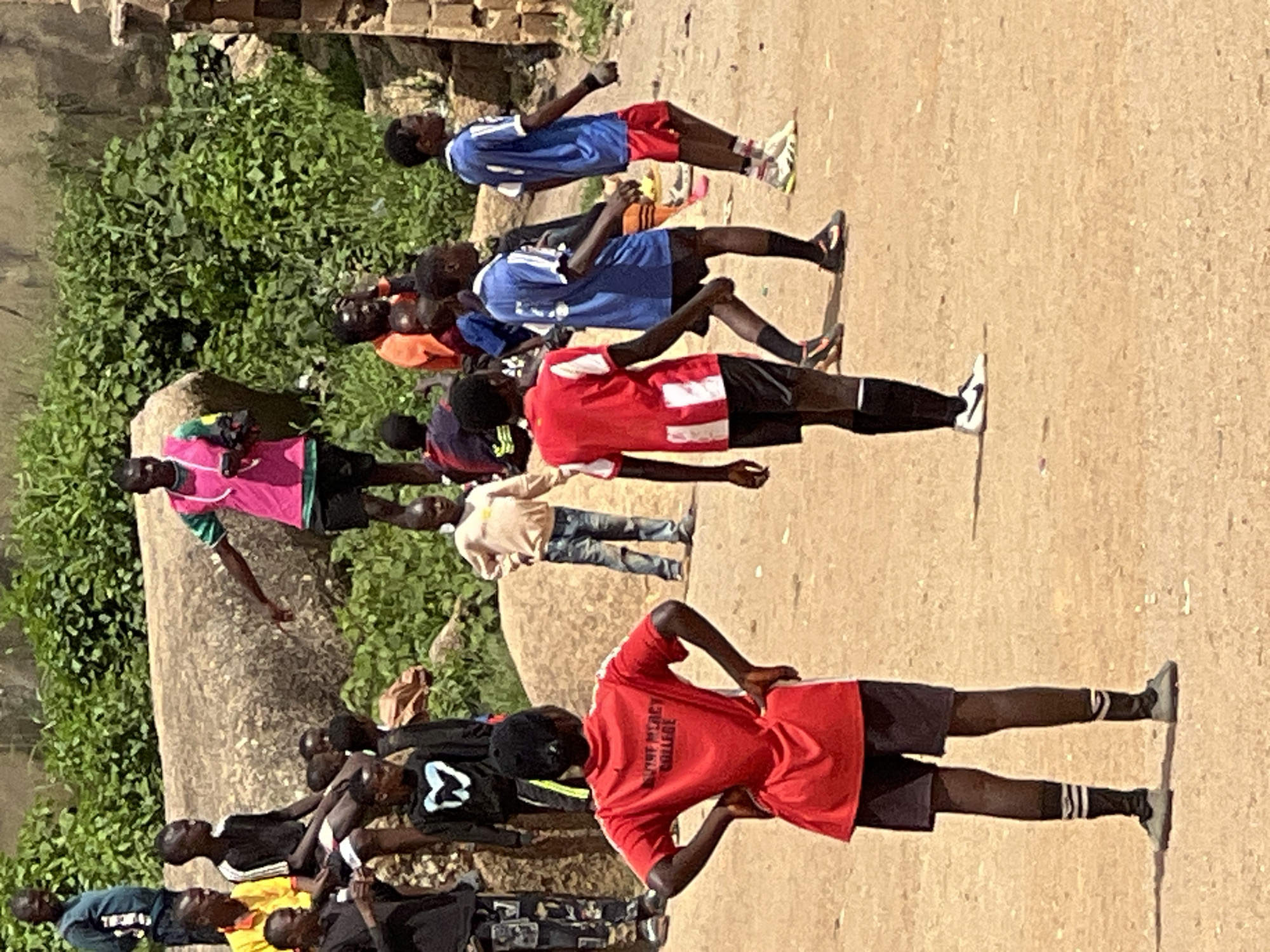 The image depicts an outdoor scene in Jos, Nigeria, likely a sports field or recreational area, given the presence of children in athletic attire. In the foreground, a child wearing a red t-shirt and black shorts with white striped socks and cleats is facing away from the camera, with their hands on their hips. Several other children are visible in the middle ground, some in blue soccer jerseys and shorts, others in casual clothing. One child in the background, wearing a bright pink vest over a green shirt, appears to be directing or observing the group. The environment is characterized by a dusty, sandy ground and a backdrop of rocky terrain with lush green vegetation, suggesting a semi-arid or sub-tropical climate typical of Jos. The overall atmosphere appears to be one of informal activity, possibly a practice session or a casual game.