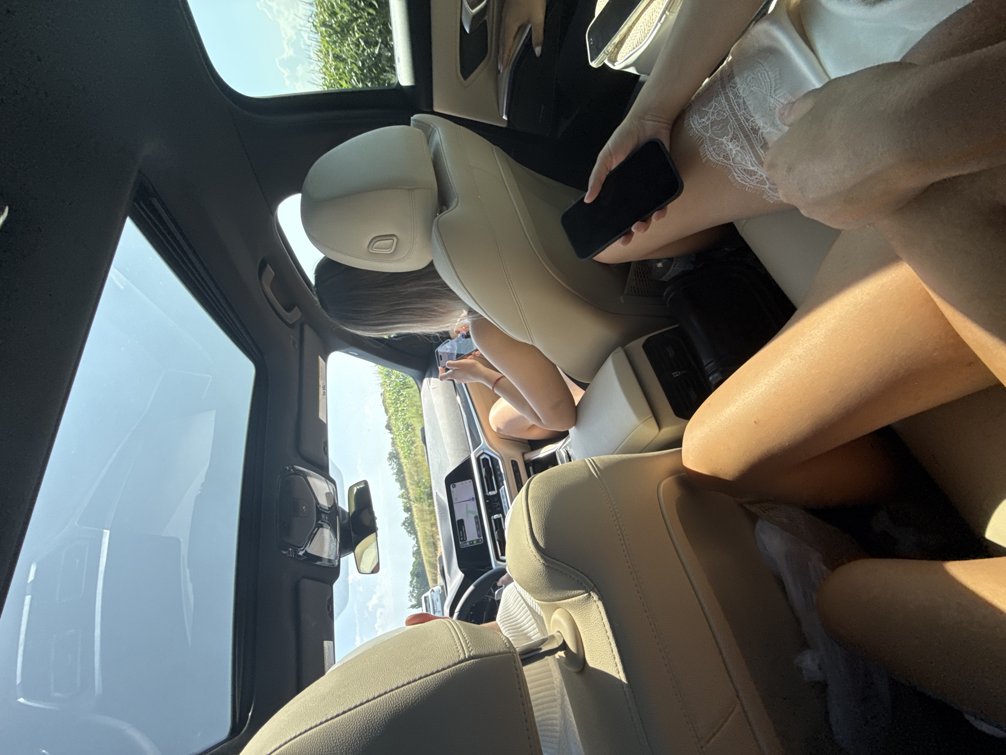 Inside a car, the camera faces forward, capturing a view of the road ahead through the sunroof and windshield. The interior is illuminated by natural light. Two individuals are visible. In the front passenger seat, a person with long blonde hair is holding a smartphone. In the rear passenger seat, another person's legs are visible, holding a smartphone. The car appears to be on a rural road, with a field of crops visible on the right side of the frame and trees lining the horizon. The sky is clear and blue with a few scattered clouds. The car's dashboard and infotainment system are also visible. It is not possible to determine the exact location of Sâmbăta de Jos, Romania from this image.