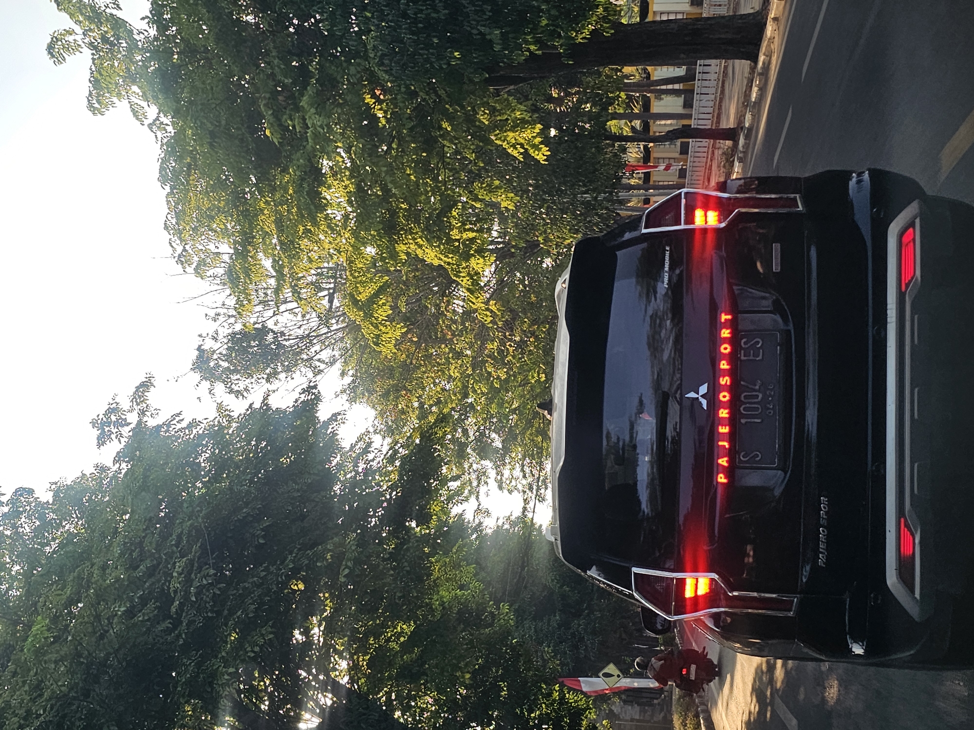 The image shows the rear of a black Mitsubishi Pajero Sport driving on a road in Tuban, Indonesia. The car's brake lights are illuminated, indicating it is slowing down or stopped. The license plate is visible, reading "S 1004 ES". The vehicle is surrounded by lush green trees that line the road, with sunlight filtering through the leaves, creating a dappled effect. A person on a scooter is visible in the left side of the frame, also on the road. In the background, there are buildings and a white fence. The sky is bright, suggesting it is daytime.