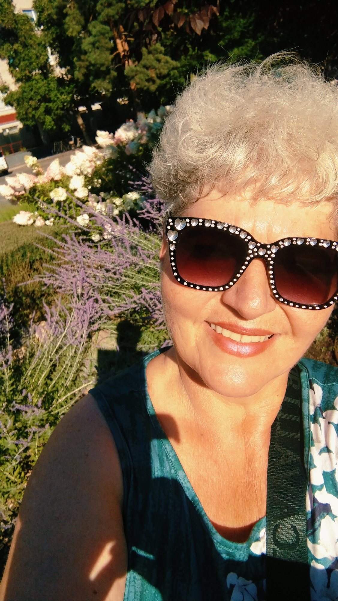 A woman with short, light-colored, curly hair and fair skin is captured in a close-up selfie. She is wearing large, black, cat-eye sunglasses adorned with rhinestones. Her lips are slightly parted, revealing her teeth, and she appears to be smiling. She is wearing a teal-colored sleeveless top with a floral pattern. The background is softly blurred, showing greenery, trees, and what appears to be pink and white flowers, suggesting an outdoor setting. The sunlight casts shadows on her face and clothing. The overall impression is that of a candid, outdoor portrait.