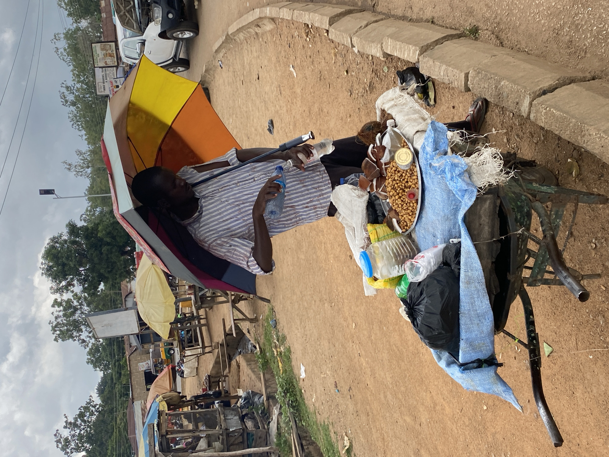 An outdoor scene in Bukuru, Nigeria, depicts a man standing under a colorful umbrella, holding a bottle of water. He appears to be a vendor, with a cart in front of him displaying various food items, including nuts, coconuts, and what seem to be packaged goods. The cart is covered with a blue tarp. In the background, there are other market stalls, vehicles, and trees, suggesting a bustling commercial area. The ground is made of red earth, and a concrete curb runs along the side of the road. The sky is overcast with clouds.