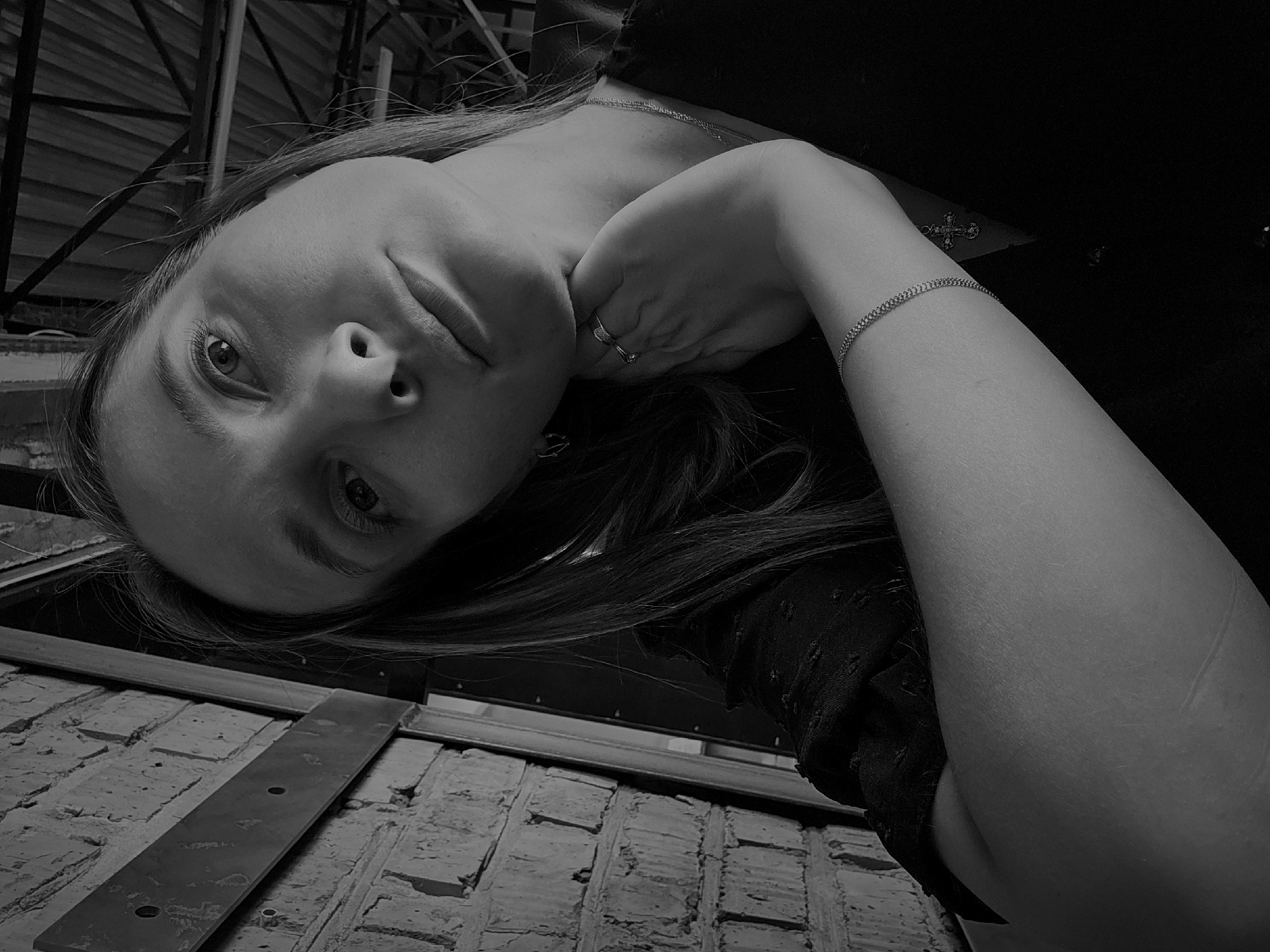 A black and white, low-angle photograph depicts a woman with long hair, a ring on her finger, and a bracelet. Her hand is positioned near her neck. She is wearing a shirt and a necklace with a cross. The background includes a brick wall and what appears to be structural metal beams. The location cannot be determined from this image alone. The city of Chaikovsky, Russia is not apparent.