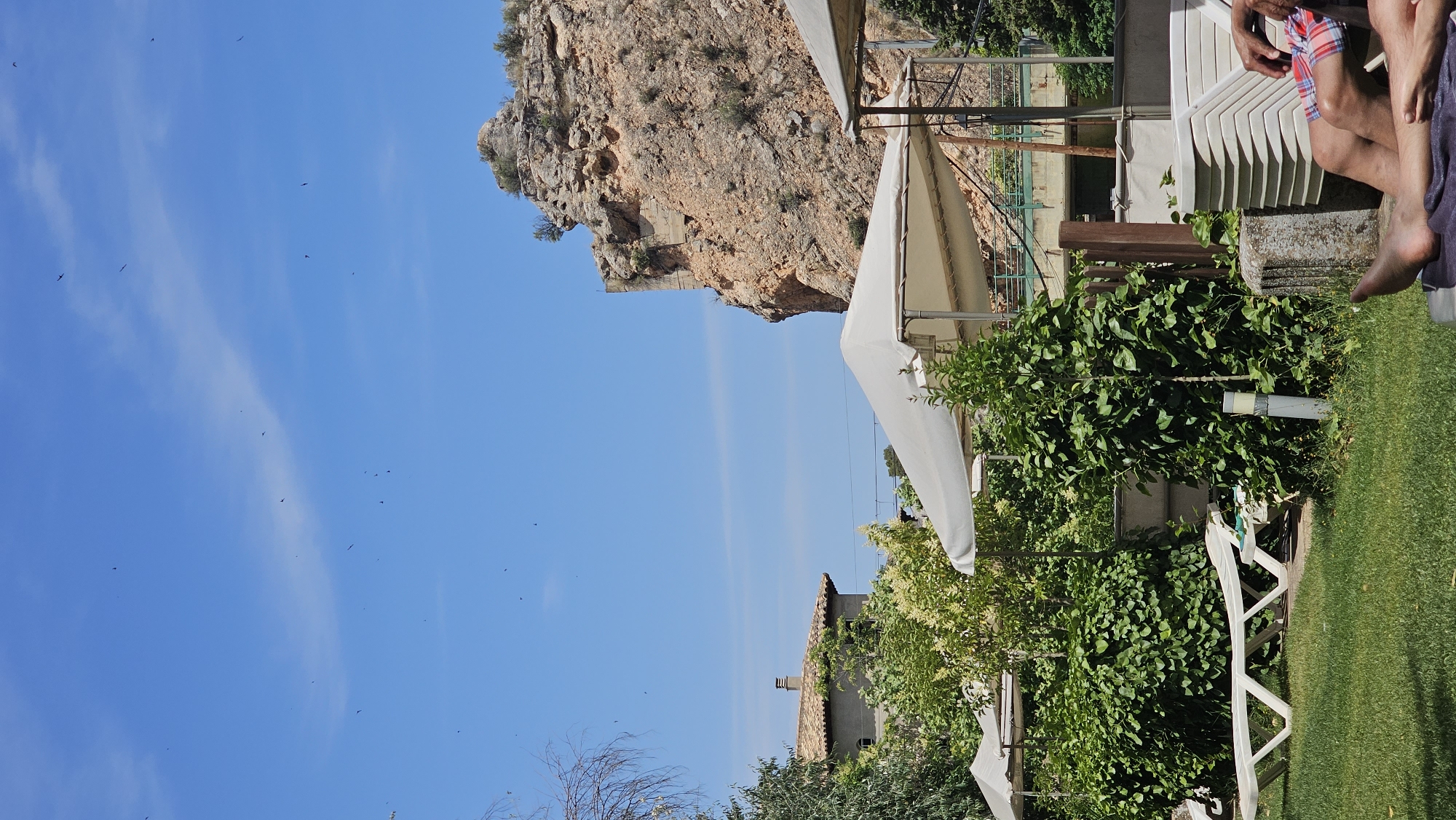 The image depicts an outdoor scene at what appears to be a resort or recreational area in Alhama de Aragón, Spain. In the foreground, a person's legs are visible, clad in plaid shorts, resting on lounge furniture. A neatly mowed green lawn occupies the lower portion of the frame, with a white plastic sun lounger positioned on it. Lush green bushes and trees are prominent throughout the mid-ground, interspersed with white canopies providing shade. To the left, a building with a tiled roof and a visible chimney can be seen behind the foliage. Dominating the background is a large, rocky cliff face with signs of past construction or stabilization. The sky is a clear, bright blue, dotted with numerous small, dark shapes that appear to be birds in flight.