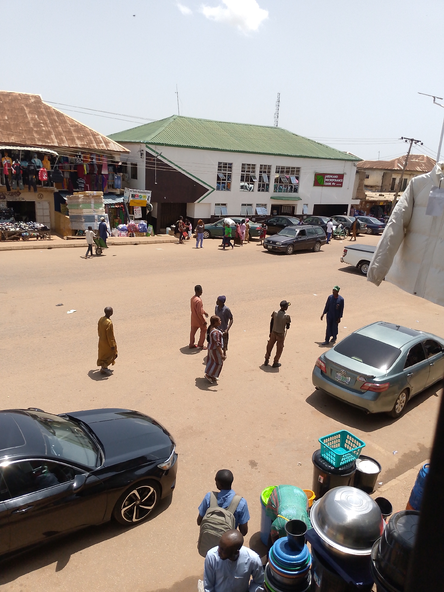 In Bukuru, Nigeria, a bustling street scene unfolds under a bright sky. Shops line one side of the road, displaying colorful textiles and merchandise. A bank building, Highland Microfinance Bank Plc, stands prominently with its green roof and white facade. People are scattered across the street, engaged in various activities: some walk, others stand in groups, and a man pushes a wheelbarrow. Several cars are parked or driving on the road, including a black sedan, a grey station wagon, and a light blue sedan. In the foreground, a collection of plastic containers and metal bowls are stacked, suggesting a marketplace or vendor stall. The ground is a dusty, earthen surface.