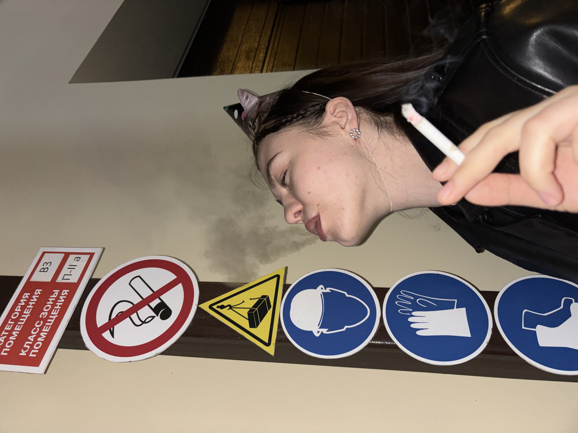 The image shows a woman in a black leather jacket, wearing cat ears, exhaling cigarette smoke near a wall. The wall is decorated with safety signs, including "No Smoking", "Caution Crane Overhead", "Wear Helmet", "Wear Gloves", and "Wear Safety Shoes". A red sign with Cyrillic text is also present. The location is Unknown, Unknown.