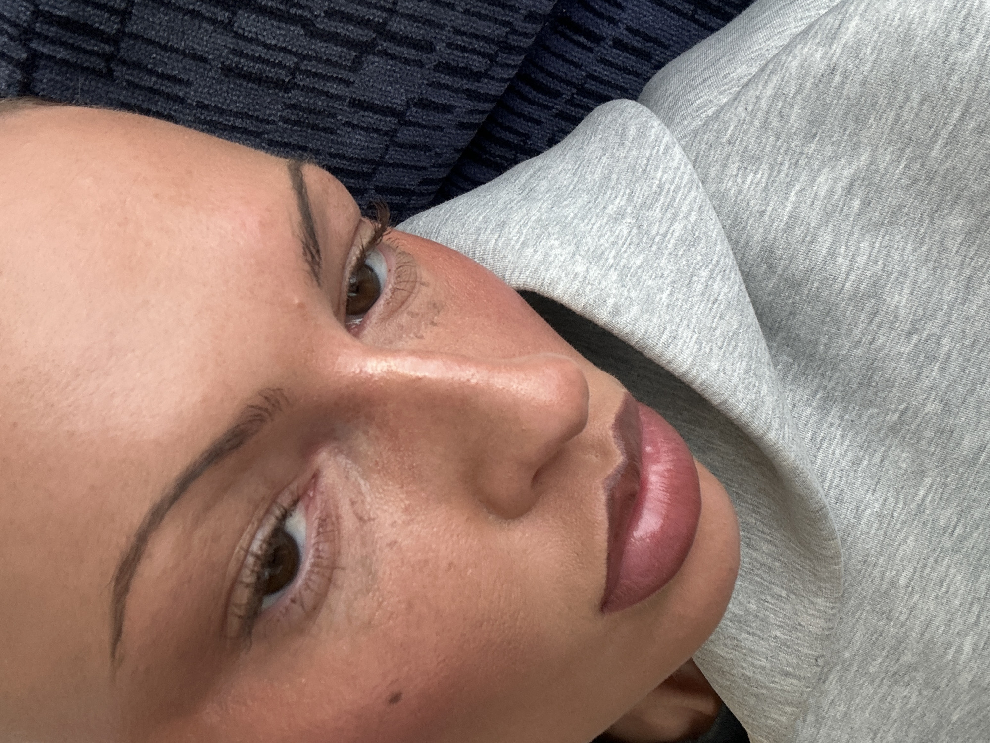 Close-up of a fair-skinned person's face tilted towards the camera, revealing the right eye, eyebrow, nose, and lips. The person has brown eyes and thin, arched eyebrows. The lips are a glossy pink color. The background is a blurry combination of dark blue textured fabric and a gray heathered fabric, likely clothing. There is no indication of location in London, United Kingdom.