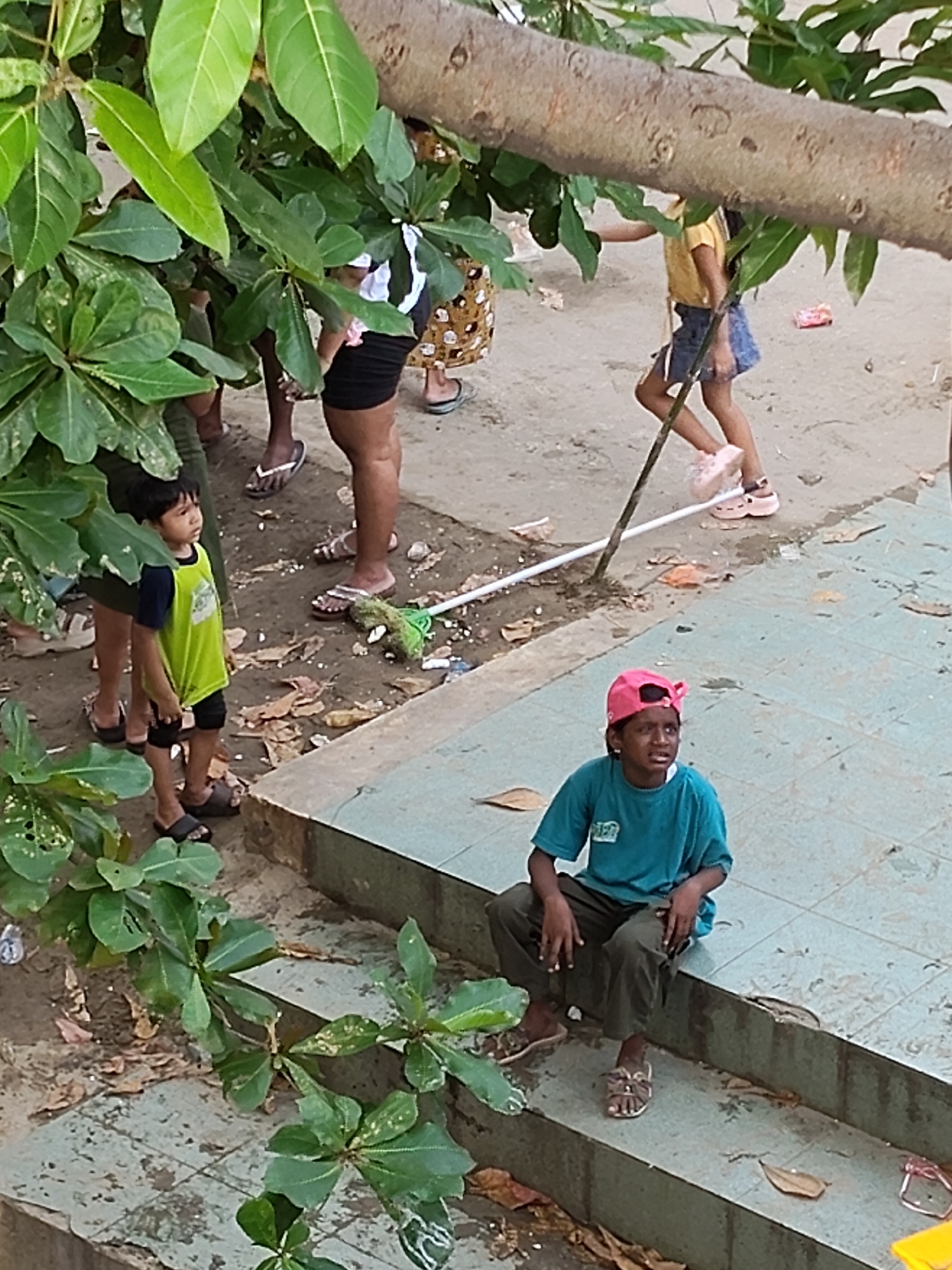 The image captures a ground-level view of a sandy area with some paved steps, possibly in a park or public space in the City of Medan, Indonesia. In the foreground, a young child wearing a bright lime green t-shirt and dark shorts stands looking to the left, partially obscured by large green leaves from an overhanging tree. To the right, another child, wearing a teal t-shirt and dark pants, sits on the edge of the paved steps, looking to the right with a pink baseball cap on their head. Several adults are visible in the background, some partially hidden by foliage. One adult is seen wearing a patterned skirt and a white top. The ground is a mix of sand and fallen leaves, with some scattered debris, including a green mop. The overall scene suggests a casual outdoor gathering or activity.