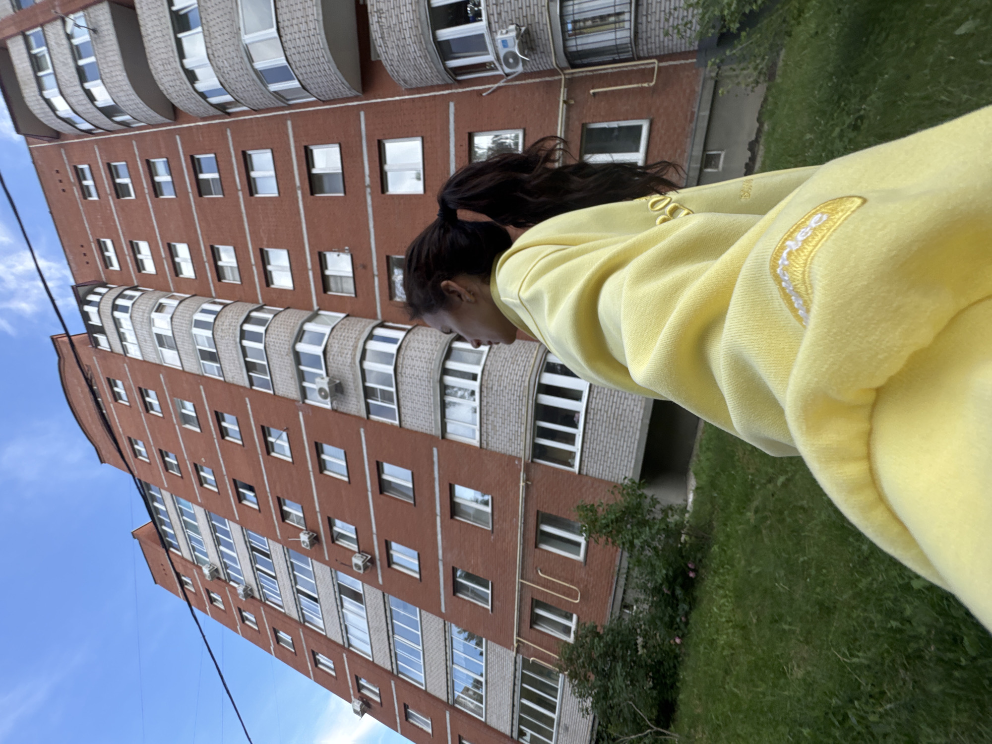 A young woman with dark hair pulled into a ponytail stands in a grassy area looking up at a tall, red brick apartment building. She is wearing a bright yellow hooded sweatshirt with some yellow embroidery on the sleeve and chest. The building has many windows and balconies and appears to be in a residential area. The sky is blue with a few wispy clouds. There is a black cable running diagonally across the upper portion of the image. The overall scene suggests a casual, outdoor setting in a Russian city.