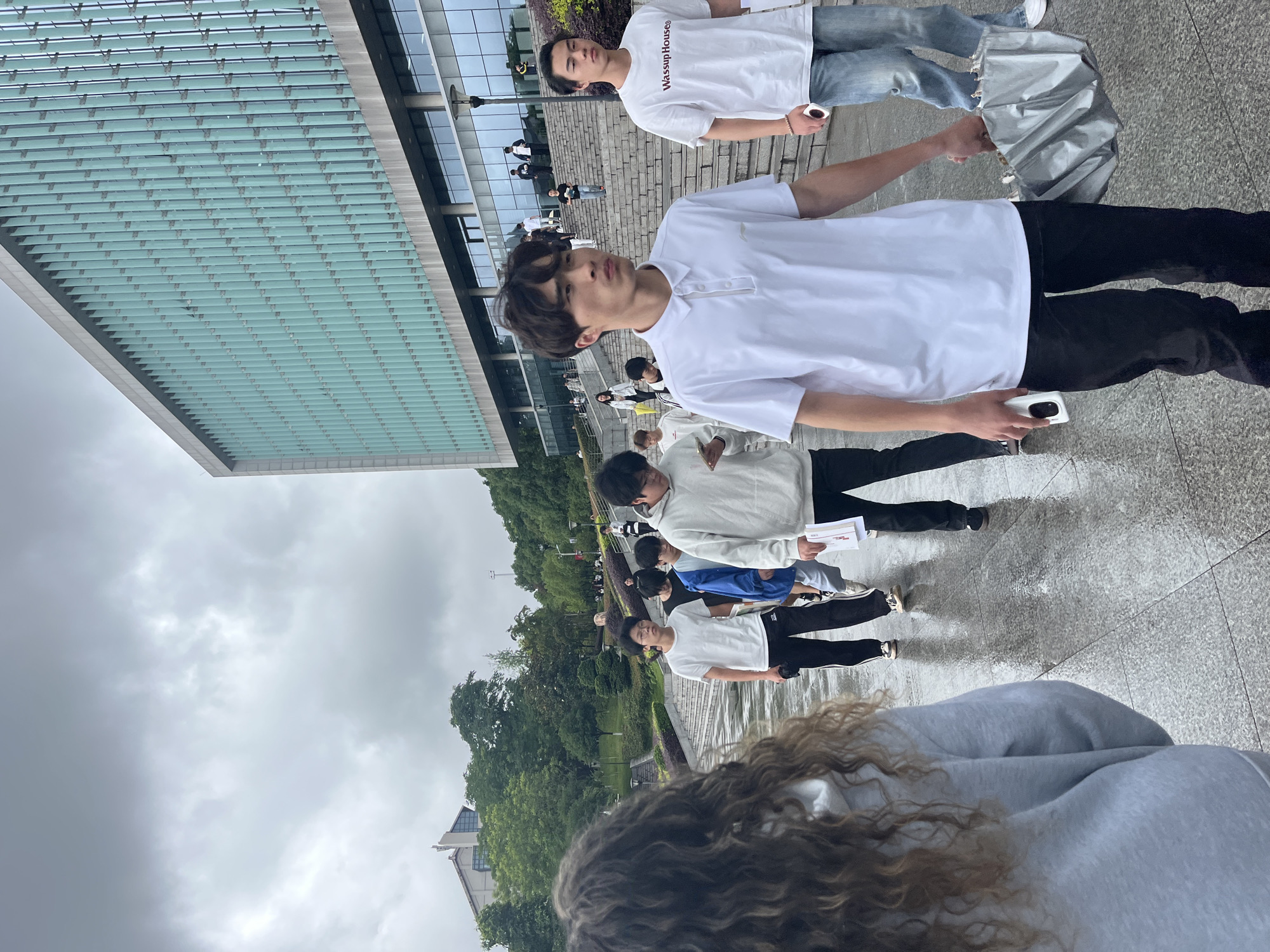 The image depicts an outdoor scene on a cloudy day, likely on a university campus in Jiangjin, China, given the architectural style of the building. In the foreground, a young woman with curly, light brown hair is seen from behind, wearing a grey hooded sweatshirt. She is facing away from the camera, walking towards a group of individuals.
A line of young men, dressed casually in t-shirts and pants, are walking up a wet, paved pathway. The person closest to the camera, a young man in a white polo shirt and black pants, is walking towards the right of the frame. To his right, another young man in a white t-shirt with the text "Wassup House" and blue jeans is also walking. The pathway is wet, indicating recent rain, and reflects the overcast sky and surrounding figures.
Behind this group, more young people are seen walking up a series of concrete steps that are bordered by landscaping. In the background, a large, modern building with a distinctive glass facade featuring vertical elements is visible. Trees and greenery are also present on the slopes leading up to the building. The overall atmosphere is that of students moving about on campus, possibly returning from or heading to classes. The weather appears to be overcast and possibly damp.