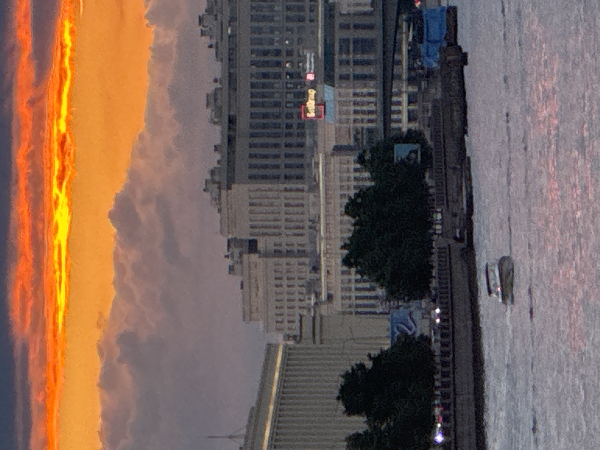 The image captures a wide shot of a city skyline at dusk, likely Saint Petersburg, Russia, given the architectural style and the presence of a large river. The sky is dominated by vibrant orange and yellow hues from the setting sun, with dramatic clouds casting shadows across the cityscape. In the foreground, a river flows, reflecting the colors of the sky. A small motorboat is navigating the water. Along the riverbank, a promenade or embankment is visible, lined with trees and what appear to be lights. Behind the trees, several large, imposing buildings with classical architecture are prominent. One building, in the mid-ground, features a prominent red neon sign that reads "SetGroup." The overall atmosphere is serene and dramatic, with the fading light creating a beautiful, painterly effect.