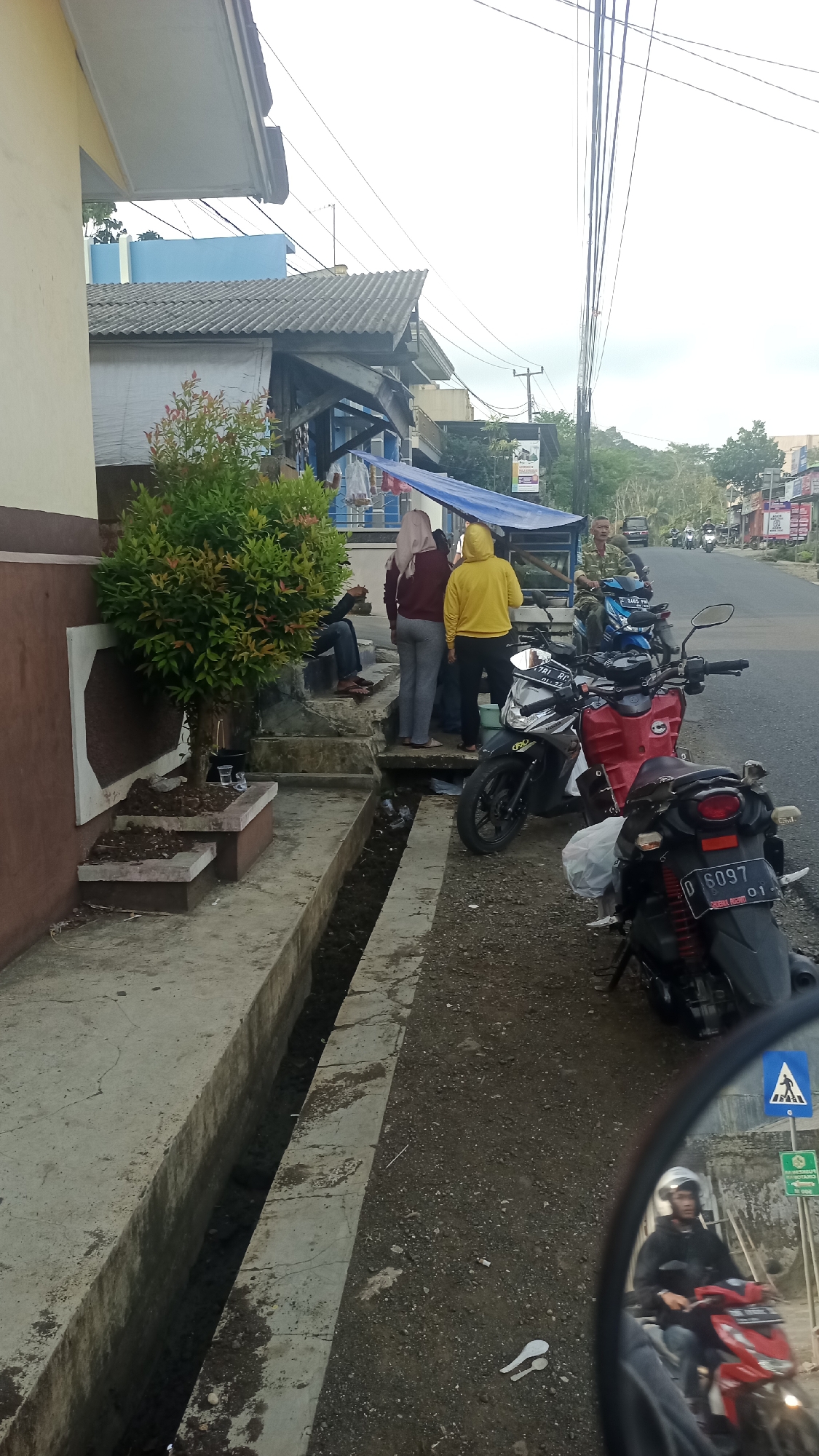 The image captures a street scene in Karyamandala, Indonesia, with a focus on a row of small shops and parked motorcycles. In the foreground, to the left, is a drainage ditch running alongside a concrete walkway. A small, ornamental tree with red-tipped leaves is planted in a tiered concrete planter near the building. Several people are standing on steps leading up to one of the shops. One person, wearing a yellow hooded jacket, is facing away from the camera, while another, in a maroon top and grey pants, is also facing away. A third person in dark clothing is seated on the steps.
Parked on the right side of the walkway are several motorcycles. A red motorcycle is prominently displayed, with a black motorcycle behind it. A third motorcycle, black and white, is parked further down the street. A man wearing a military-style uniform is seated on a blue motorcycle, also parked on the right. In the background, the street continues with more buildings and trees lining the roadside. The sky is overcast. The reflection in what appears to be a motorcycle's side mirror shows a person riding a red motorcycle and wearing a helmet, with a pedestrian crossing sign visible behind them.