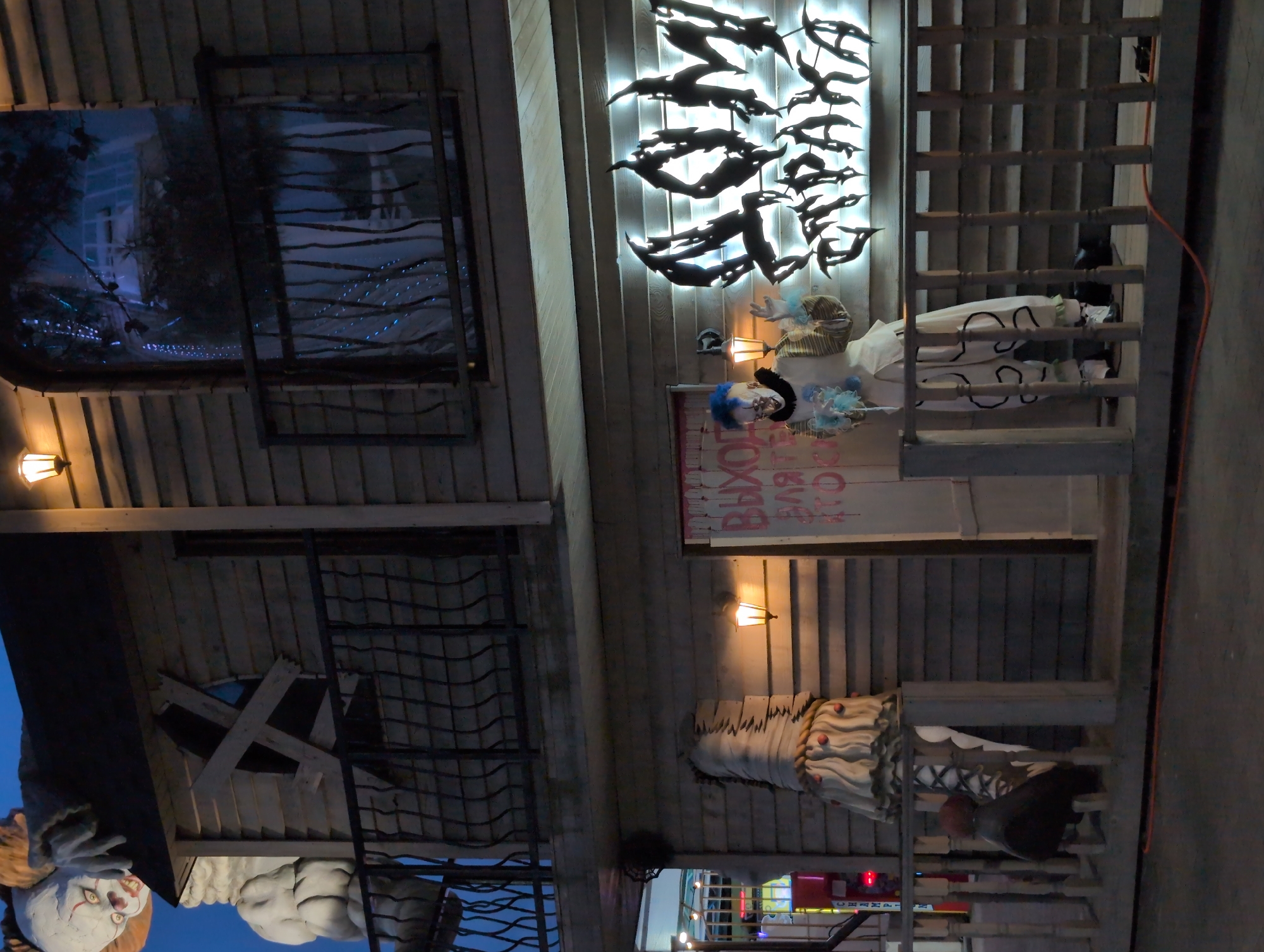 An outdoor scene at night depicts a two-story wooden structure that appears to be a haunted attraction or themed establishment. The top portion of the structure features a large, white, and red clown figure with an unsettling smile, leaning over the roof. A window on the upper level is boarded up with wooden planks in a cross shape. A balcony with a dark metal railing runs along the second floor.
The lower level has a prominent sign with stylized, illuminated Cyrillic lettering that reads "ДОМ СТРАХА" (Dom Strakha), meaning "House of Fear." In front of the sign and to the left of an entrance, a person dressed as a clown with blue hair, pale makeup, and a patterned costume stands holding a bouquet of blue flowers. This clown has a menacing expression.
To the left of the clown, a portion of another figure, possibly a mannequin dressed in white wrappings and a layered skirt with red accents, is visible. A lit lamp is mounted on the wooden wall near the clown and the entrance. The entrance door has red spray-painted Cyrillic text on it. A wooden railing with vertical posts and horizontal slats extends across the front of the lower level. In the background, to the far left, part of a colorful arcade or game machine is visible. The ground in the foreground is paved, and an orange cable runs along the ground in front of the railing. The overall atmosphere is dark and spooky, consistent with a Halloween or horror-themed event. The location is identified as Gelendzhik, Russia.