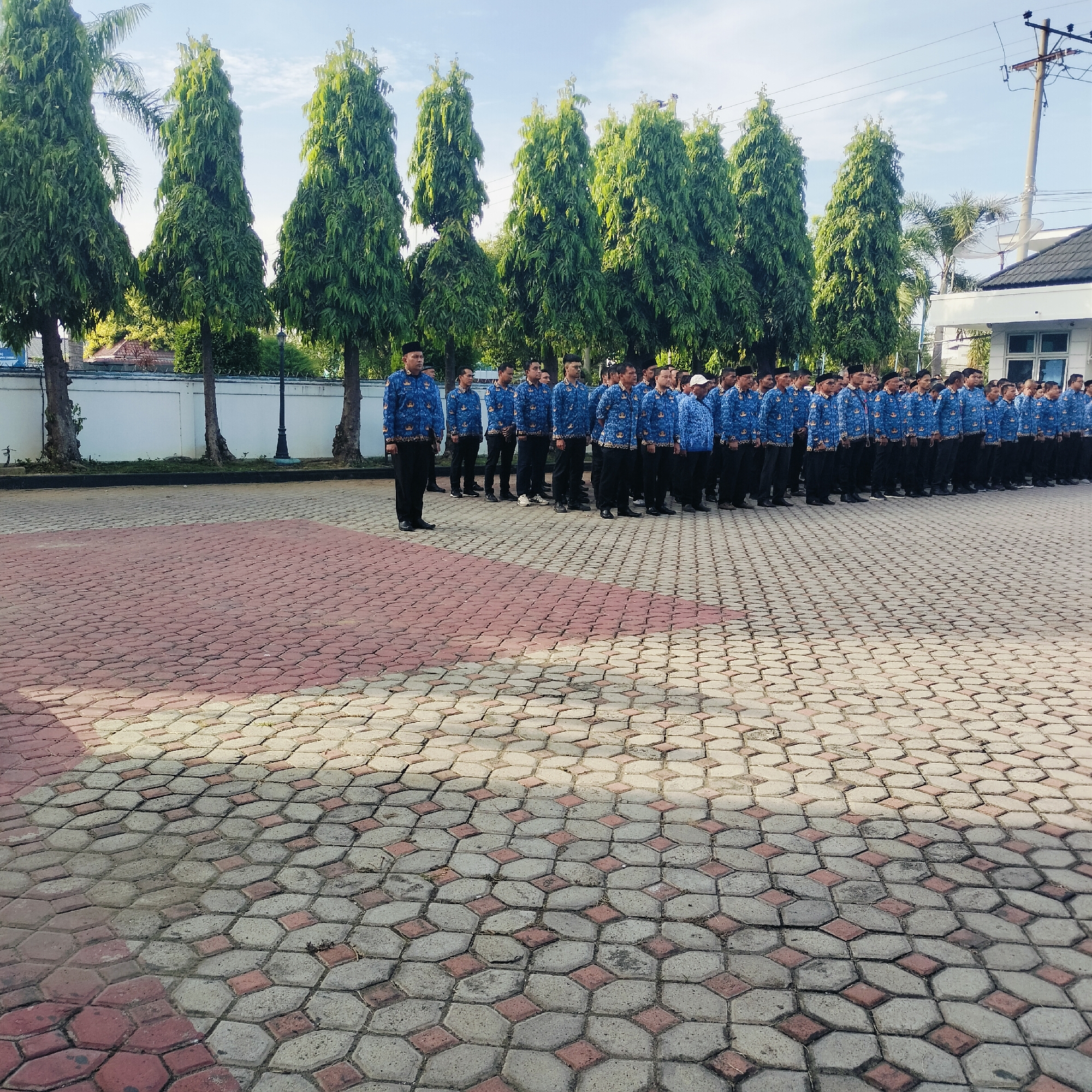 A daytime scene shows a large group of uniformed men standing in formation outdoors in Lhokseumawe, Indonesia. They are wearing blue, patterned shirts and dark trousers. The group is arranged in neat rows on a paved area with a patterned brick surface. Behind them are tall, thin trees lined up along a wall. In the background, there are buildings, power lines, and a clear sky. The light suggests it is midday or early afternoon.