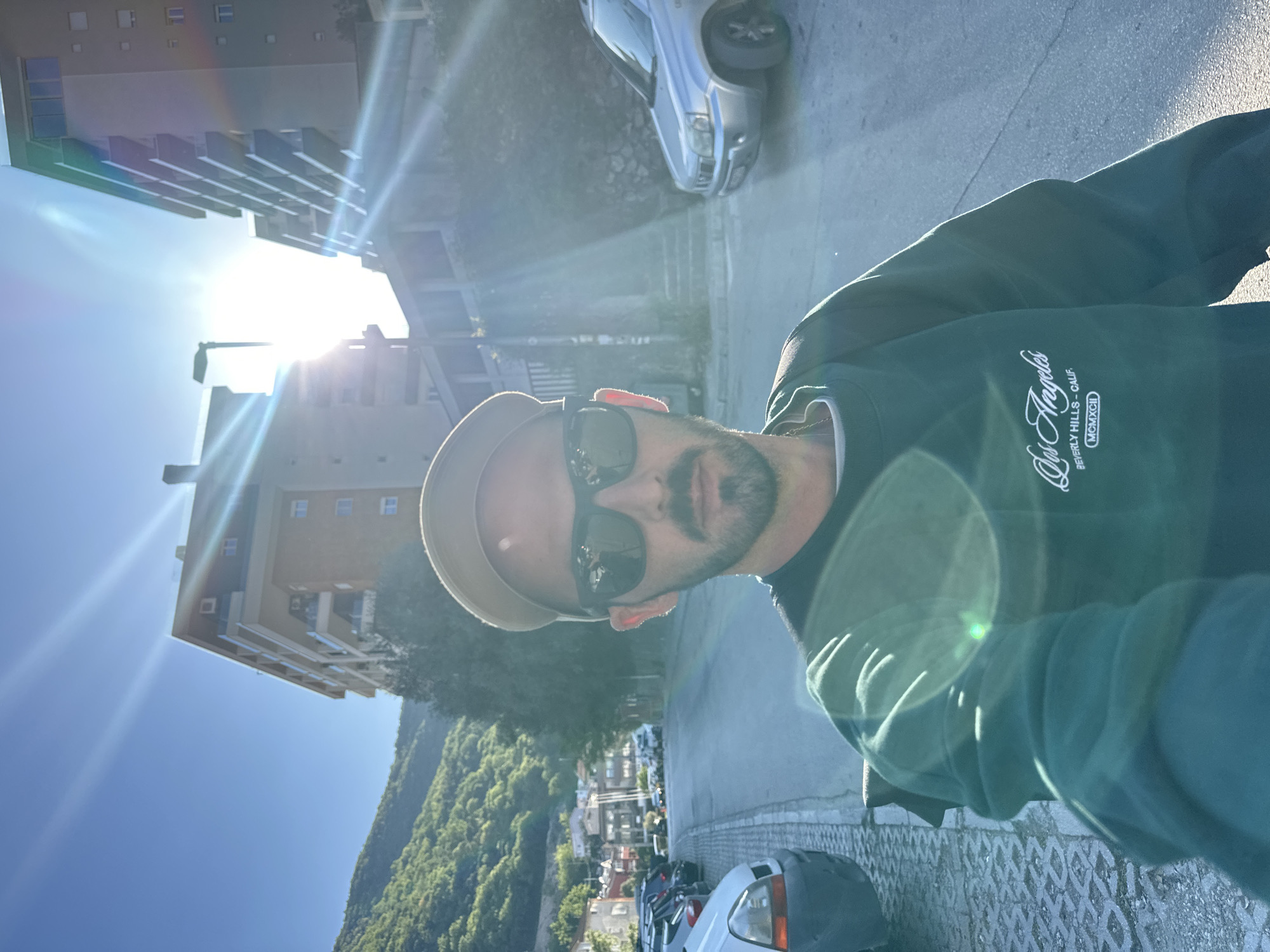 A selfie taken on a sunny day shows a man wearing a cap, sunglasses, and a dark green sweatshirt with "Los Angeles Beverly Hills" inscribed on it. He stands on a paved road or sidewalk. Buildings, a street with parked cars, and a green, mountainous landscape can be seen in the background. The location is likely Budva, Montenegro, due to the architectural style of the buildings and the surrounding mountain scenery.