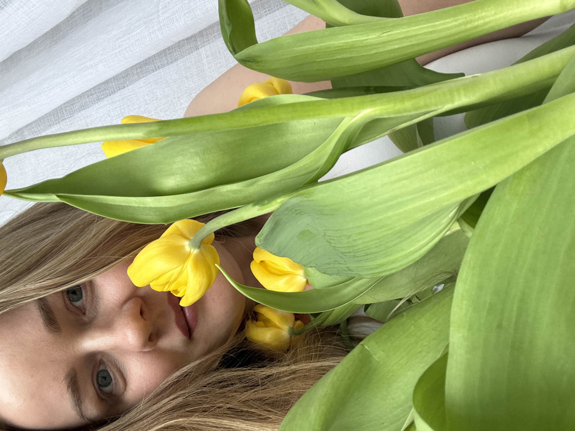 A young woman with light brown hair and gray eyes lies on a white fabric surface. She is partially obscured by a bouquet of yellow tulips with green leaves. The setting appears to be indoors.