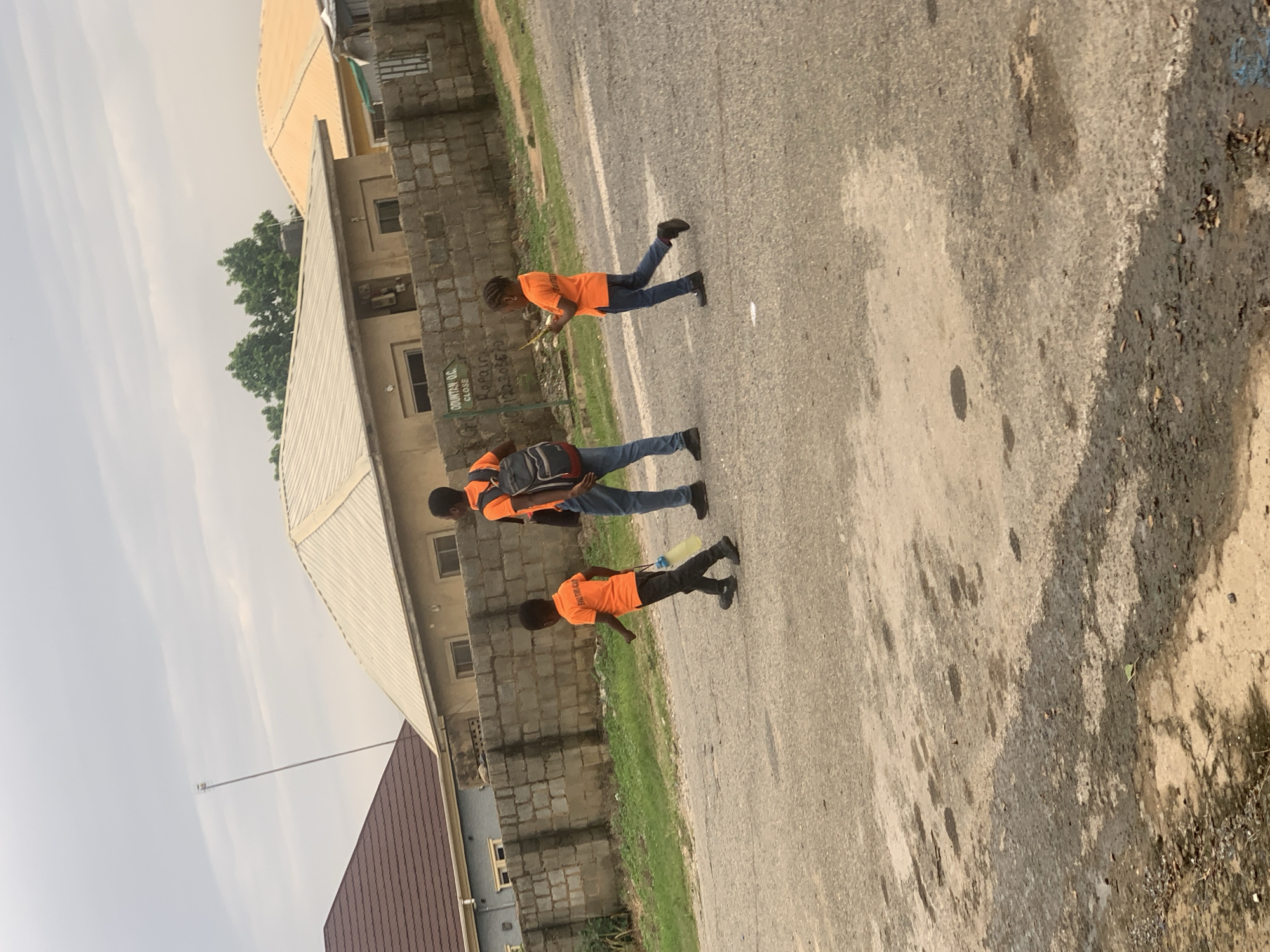 The image depicts an outdoor scene in Abuja, Nigeria, on a cloudy day. Three individuals are walking on a paved road. The person furthest to the left is a young boy wearing an orange t-shirt and dark pants, carrying a light-colored water bottle. In the center, a slightly older boy or young man, also in an orange t-shirt and blue jeans, carries a dark backpack. To the right, a young girl, wearing an orange t-shirt and blue jeans, holds a yellow object. They are walking away from the viewer, towards the right side of the frame.
Behind the individuals is a low, grey brick wall. Beyond the wall, there are several buildings with light-colored, sloped roofs. One building has a sign with text that is partially obscured but appears to include "COUNTAN O.C. CLOSE" and "REPAIRS". The environment suggests a residential or developing area within the city. The road surface is asphalt with visible patches of wetness and some debris. In the background, trees are visible above the wall, and a tall, thin metal pole with a small structure at the top extends upwards into the sky. The overall lighting indicates overcast conditions.