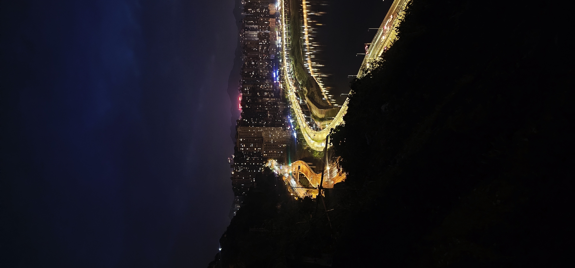The image shows a nighttime view of Yuzhong District, China. Buildings are illuminated with yellow and white lights, with a red light showing in the distance. The sky is dark, and a body of water is near the light trails. Vegetation is visible in the foreground. The image is oriented sideways.
