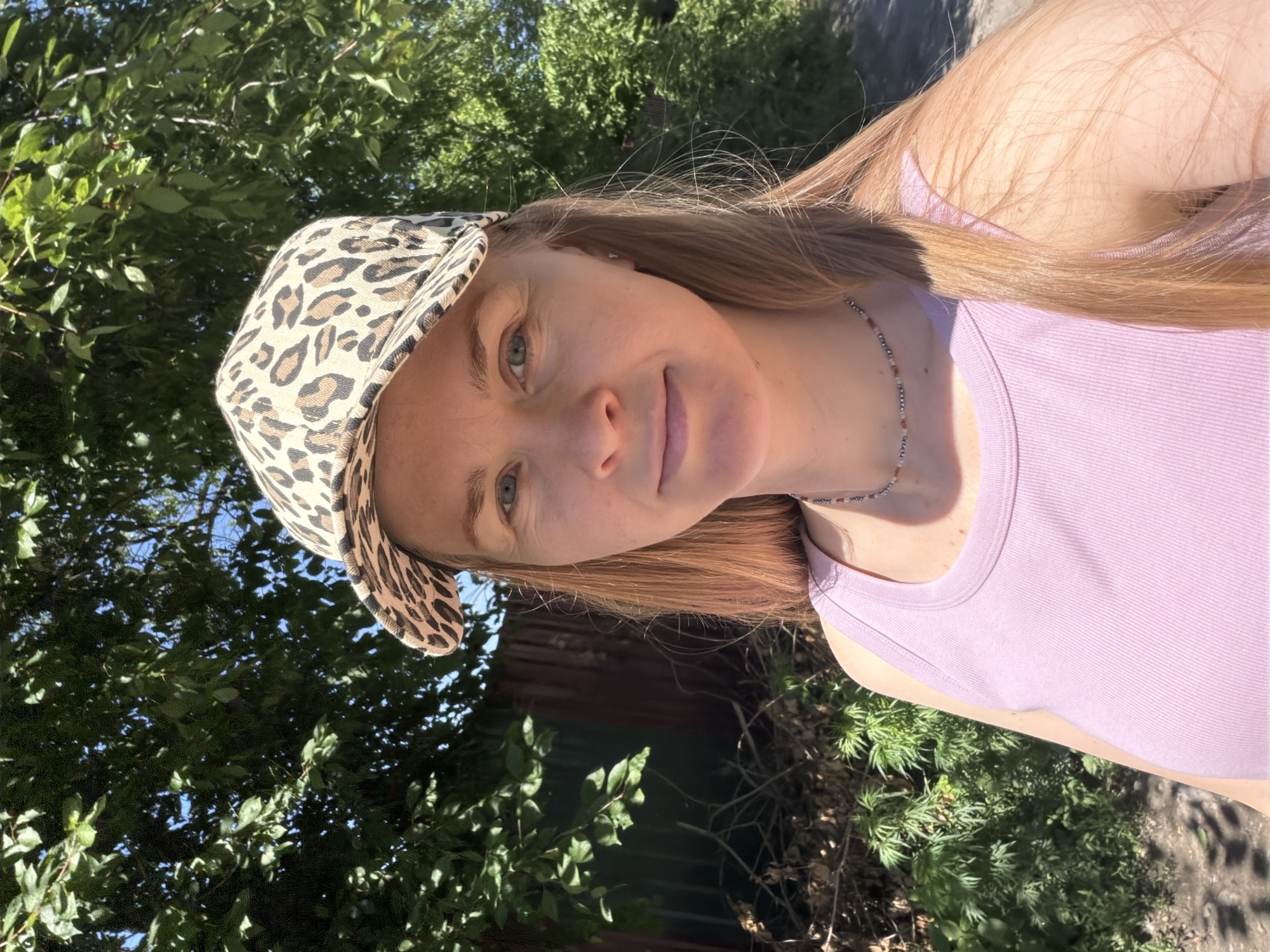 This media file is a portrait of a young woman, likely taken outdoors in a natural setting. She is wearing a leopard print baseball cap and a light purple tank top. A delicate beaded necklace adorns her neck. Her long, light brown hair is visible, framing her face. The background consists of lush green foliage, with sunlight filtering through the leaves, suggesting a bright, sunny day. The scene appears casual and relaxed, with the woman's head tilted slightly and a gentle smile on her face, conveying a sense of contentment. The overall impression is one of a warm, pleasant day spent outdoors. The location context is Khabarovsk, Russia. There is no visible text in the image.