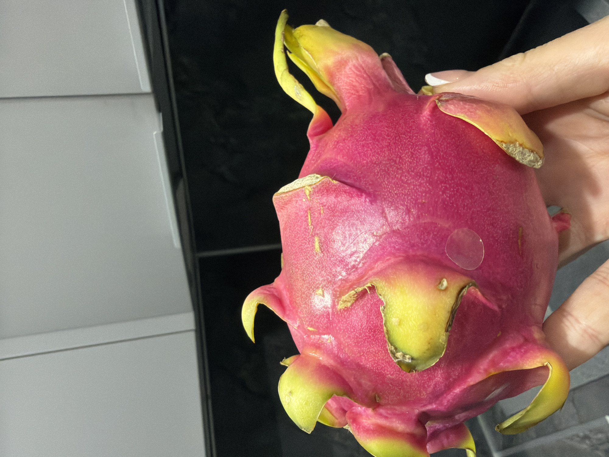 A dragon fruit is held in a person's hand. The fruit is bright pink with yellow and green scales. A clear sticker is visible on the surface of the fruit. The background is a blurry mix of black and white surfaces. The location is unknown.