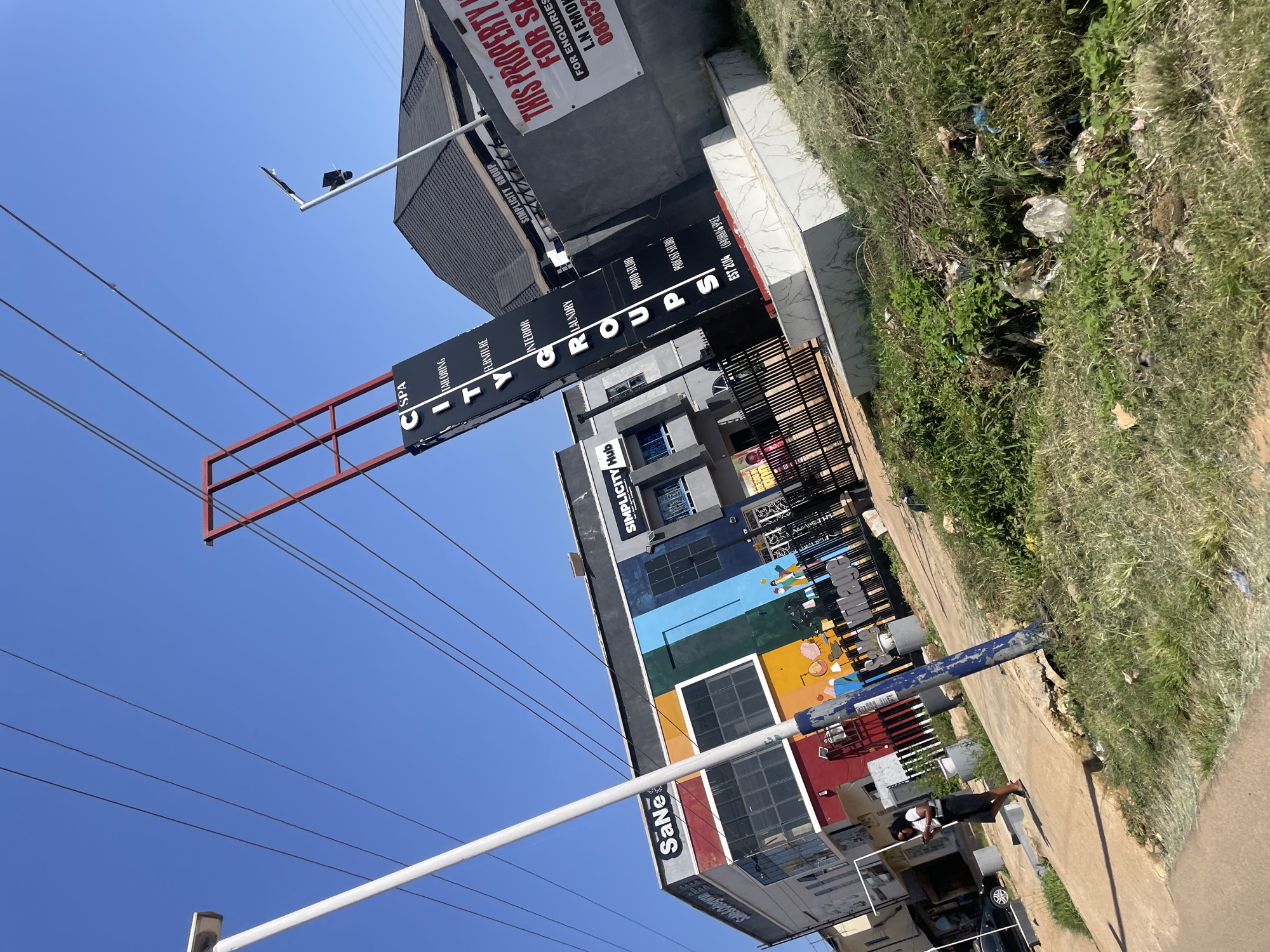 The image shows a street scene in Gura Top, Nigeria. A large black sign with red trim stands vertically, displaying "CITY GROUPS" in white letters and various businesses such as "SPA," "TAILORING," "FURNITURE," "INTERIOR," "LAUNDRY," "PHOTO STUDIO," "PODCAST STUDIO," "EST 2016," and "CO-WORKING SPACE." To the left of the sign is a building with a colorful facade featuring red, yellow, green, blue, and black panels. The building has large windows and the text "SaNe" and "SaNe & Obanwu" are visible. To the right of the sign, a gray wall displays a "FOR SALE" sign with text that reads "THIS PROPERTY IS FOR SALE FOR ENQUIRIES CALL L.N EMOH 08037007962."
In the foreground, a woman in a white shirt and black skirt is walking on a concrete sidewalk, looking at her phone. A black car is parked on the left side of the street. The environment appears to be a commercial area with buildings, a sidewalk, and a road. There is overgrown grass and scattered litter on the right side of the image, suggesting an unkempt area. Power lines are visible across the clear blue sky. The overall scene depicts an urban environment under daylight.