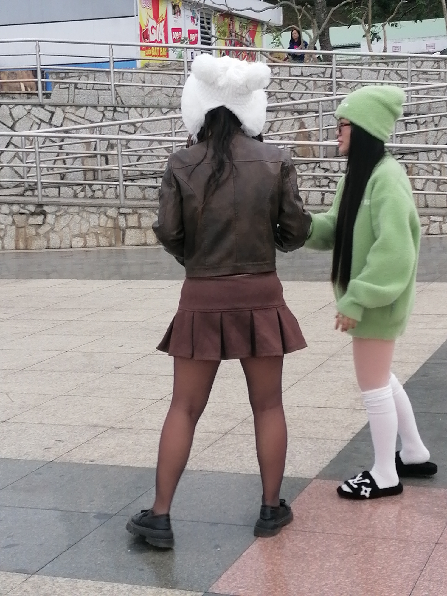 In Da Lat, Vietnam, two young women stand on a paved plaza. The woman on the left wears a white fluffy hat, a dark brown leather jacket, a brown pleated skirt, and black tights and shoes. The woman on the right, facing away from the viewer, wears a green beanie, a green oversized sweater, and white knee-high socks with black slides featuring a white LV logo. She has dark hair that extends down her back. The plaza is paved with square tiles in shades of gray and pink. Behind the women, a tiered stone structure with metal railings rises, topped by trees. A building with a brightly colored facade, featuring red and yellow signage, is visible in the background. The overall atmosphere is casual, and the women appear to be engaged in a conversation or interaction.