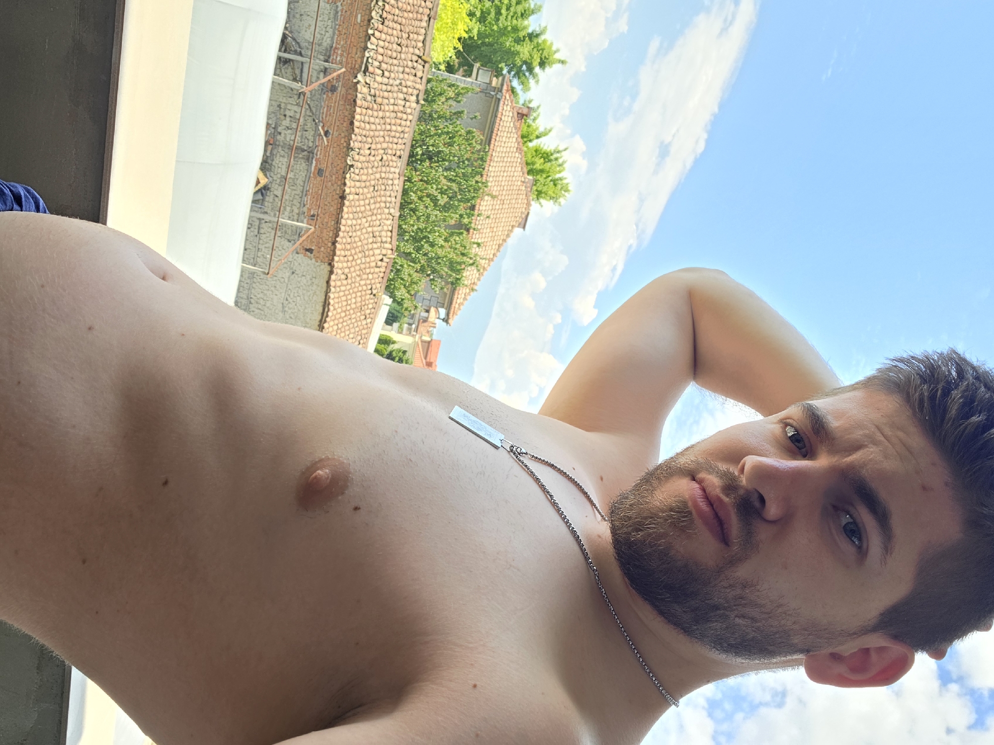 A shirtless male, appearing to be in his 20s or 30s, with a dark beard and short dark hair, is depicted looking directly at the camera from a low angle. His right arm is raised with his hand near the back of his head. He wears a silver-colored chain necklace with a rectangular pendant. The visible portion of his torso shows fair skin and some body hair. A glimpse of blue fabric is visible at the bottom left.
The background reveals an outdoor scene in Yagodovo, Bulgaria, under a clear blue sky with scattered white clouds. In the middle ground, a two-story building with a light-colored facade and a distinctive orange-tiled gabled roof is partially obscured by green trees and bushes. Below this, an older structure with a similar orange-tiled roof, exposed brickwork, and what appears to be scaffolding or structural supports is visible. Further in the distance, other residential buildings with orange roofs are partially visible, consistent with a town or village environment. The immediate foreground shows a dark, flat surface, possibly a balcony railing, behind which a white opaque barrier or wall is present. The scene is brightly illuminated by natural sunlight.