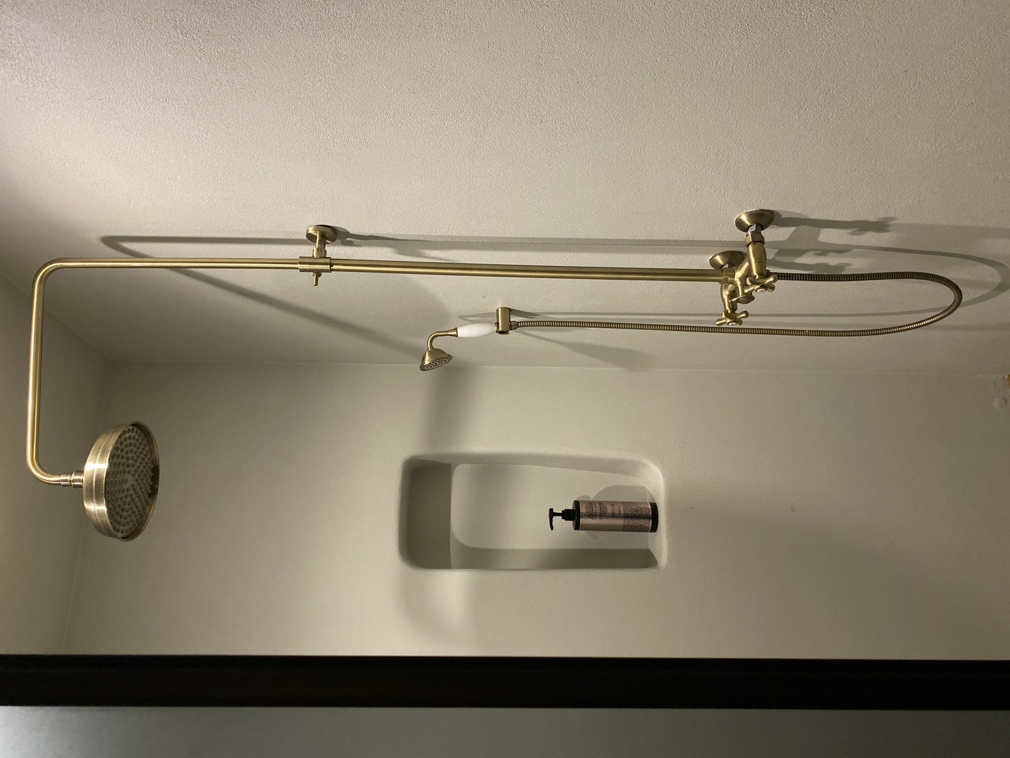 A shower stall with a brass shower fixture, including a fixed shower head and a handheld shower head, and a recessed shelf with a bottle of shampoo. The scene is likely inside a bathroom, but there is no other information to confirm its location within the city of Izhevsk, Russia.