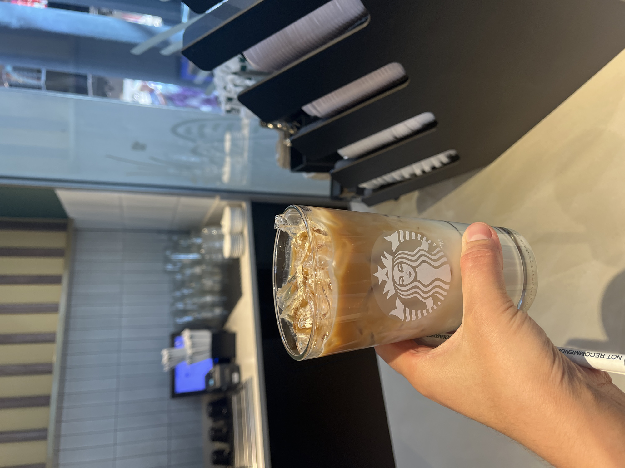 A hand holds a clear glass of iced coffee with the Starbucks logo visible. The coffee is light brown with ice cubes and a layer of milk at the bottom. The background shows a counter with a blue monitor, stacks of paper cups in a black holder, and tiled walls. The scene appears to be inside a coffee shop.