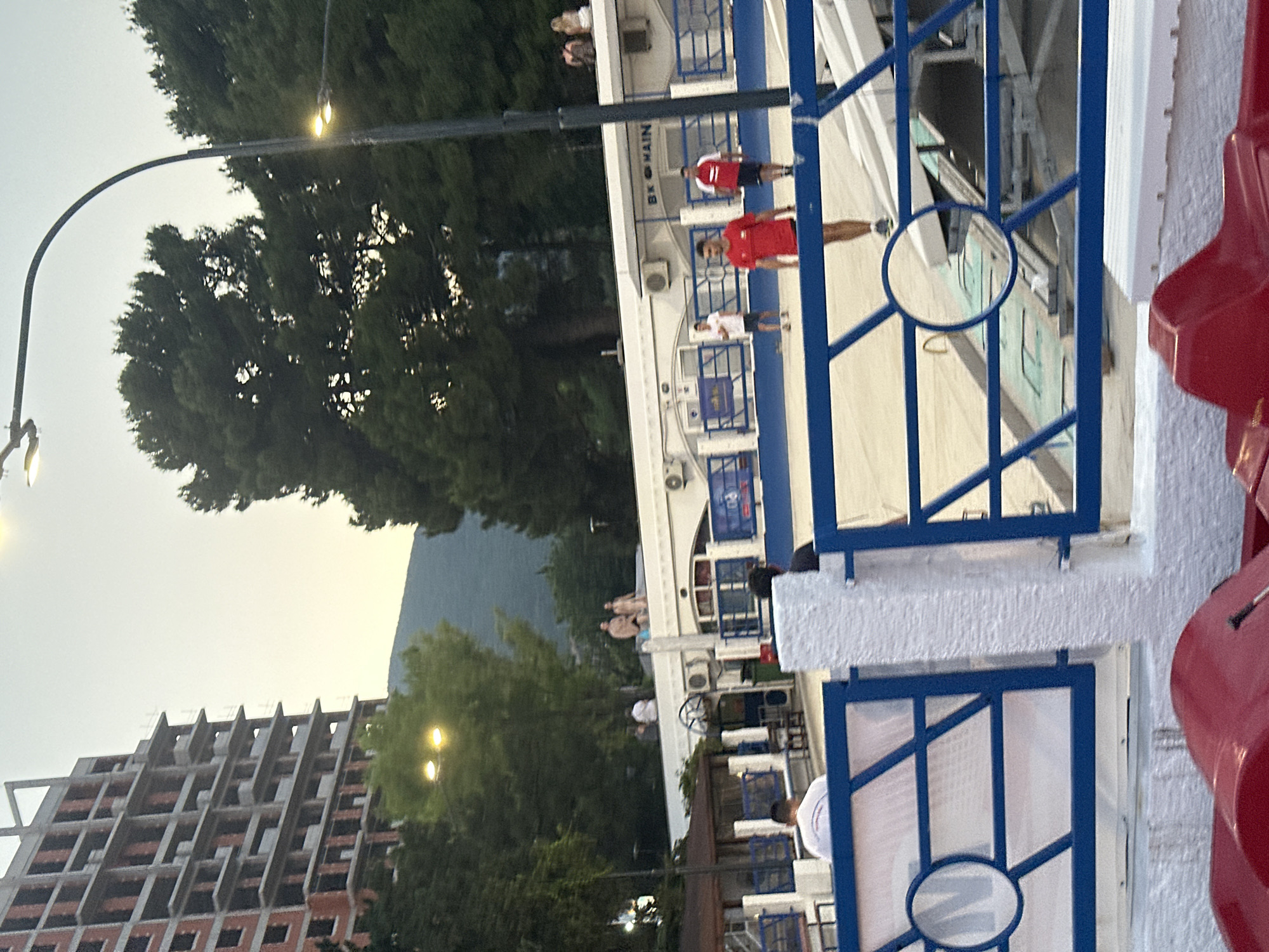 The image depicts a scene in Budva, Montenegro, likely at a recreational area or complex. In the foreground, red plastic structures, possibly parts of paddle boats or seating, are visible. Behind this, a white, textured pillar separates two blue metal fences with a circular design element. Beyond the fence, a blue and white barrier encloses a paved area where several people are engaged in activities or spectating.
In the middle ground, a white building with blue trim and air conditioning units is situated. A sign on this building reads "BK MAIN". Several individuals are present, including a man in a red shirt and shorts, another in a white shirt and shorts, and a third in a red and white shirt and dark shorts. A fourth man in a white shirt is partially visible in the background. On top of the white building, four people, two couples, are standing, their attire suggesting they are casually dressed.
The background features tall, lush green trees, obscuring a mountainous landscape in the far distance. The sky is overcast with a pale, diffused light, indicating either early morning or late afternoon. To the left, a partially constructed building, a multi-story concrete structure with exposed brickwork and unfinished floors, stands prominently. Streetlights with illuminated bulbs are visible among the trees.