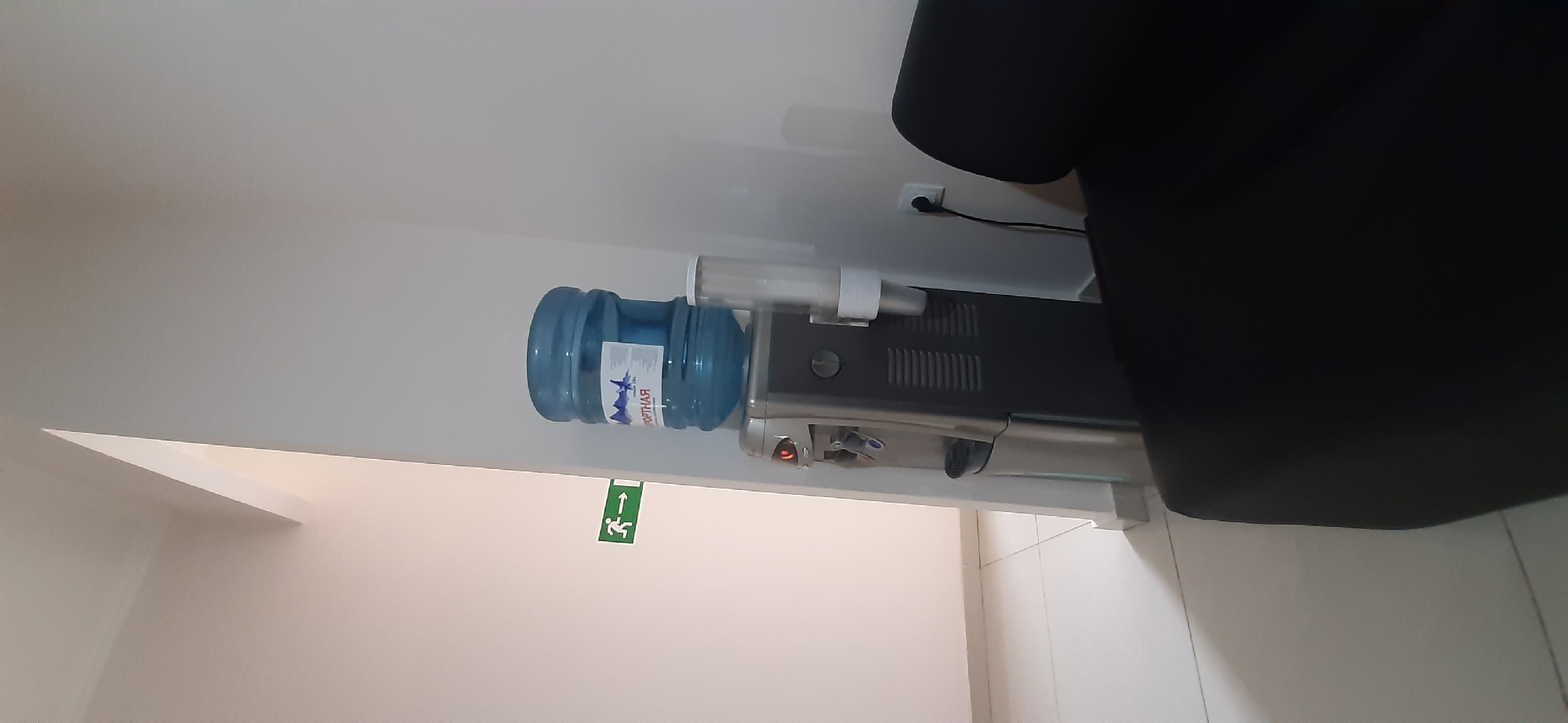 The image depicts the interior of a building, likely an office or public space, with a focus on a water cooler. The water cooler is a metallic and dark gray appliance with a large blue water bottle on top. To the left of the water cooler, a green exit sign with a white running figure and arrow is visible on a white wall. The floor appears to be tiled with white tiles. In the foreground, a portion of a black upholstered piece of furniture, possibly a sofa or chair, is visible. The overall lighting suggests an indoor environment. The scene is devoid of people.