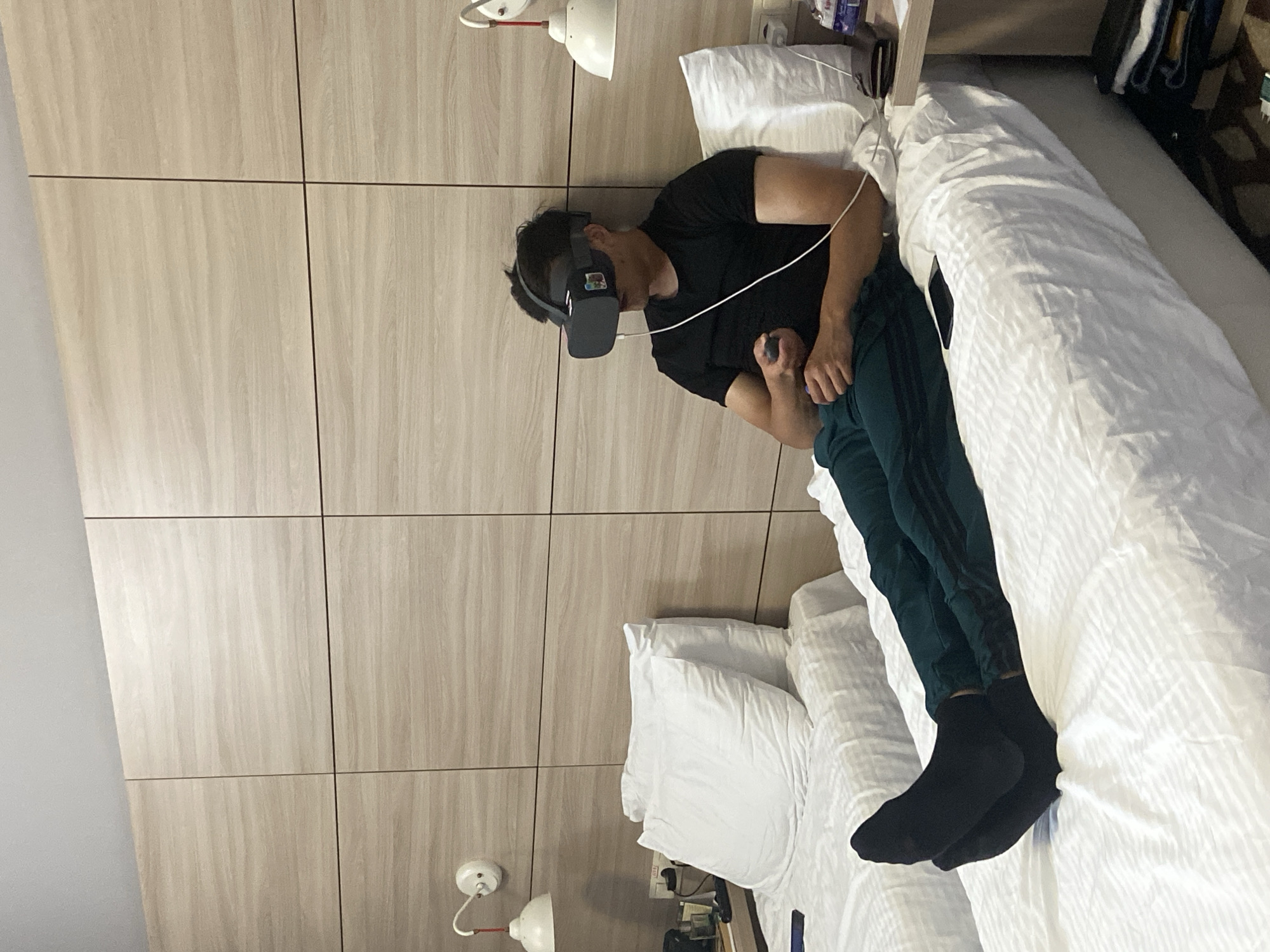 A man is lying on a bed, wearing a virtual reality headset and holding a controller. He is wearing a black t-shirt, dark green pants with three black stripes, and black socks. The bed has white linens and several pillows. The room has a light-colored wood panel wall with a light fixture on the wall. The image does not contain information necessary to confirm a location.