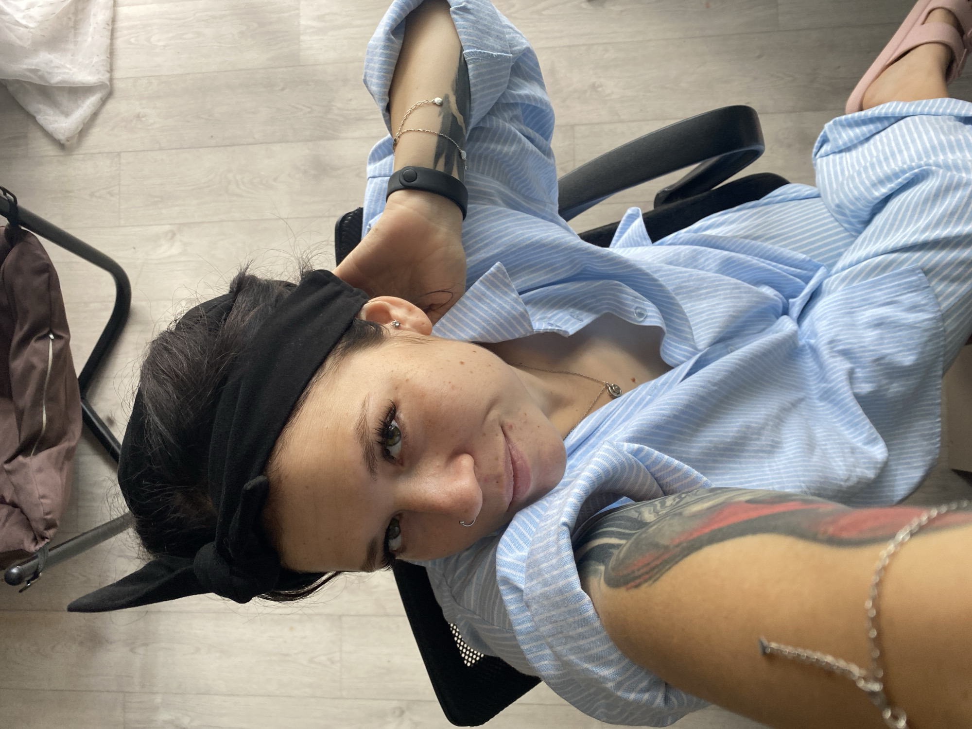 The image shows a woman sitting in a black office chair, facing the camera. She has a black headband with a bow on her head, and her dark hair is pulled back. She is wearing a light blue and white striped button-down shirt. She has a nose ring and an earring in her left ear. She has tattoos on her left arm and leg. She is wearing a bracelet on her left wrist and another piece of jewelry on her right arm. Her right hand is resting on the back of her head. Her left arm is extended towards the camera, and a silver bracelet with charms is visible on her forearm. Her legs are crossed, and she is wearing pink slip-on shoes. The background appears to be an indoor setting with light-colored wooden flooring. There is a portion of a dark brown bag or cushion and part of a metal frame of what appears to be furniture in the upper left of the image. A white piece of fabric, possibly lace or a tablecloth, is visible in the upper right corner. There are no discernible landmarks or indicators that would allow for precise location identification within the city of Chaikovsky, Russia. The scene is a casual indoor portrait.