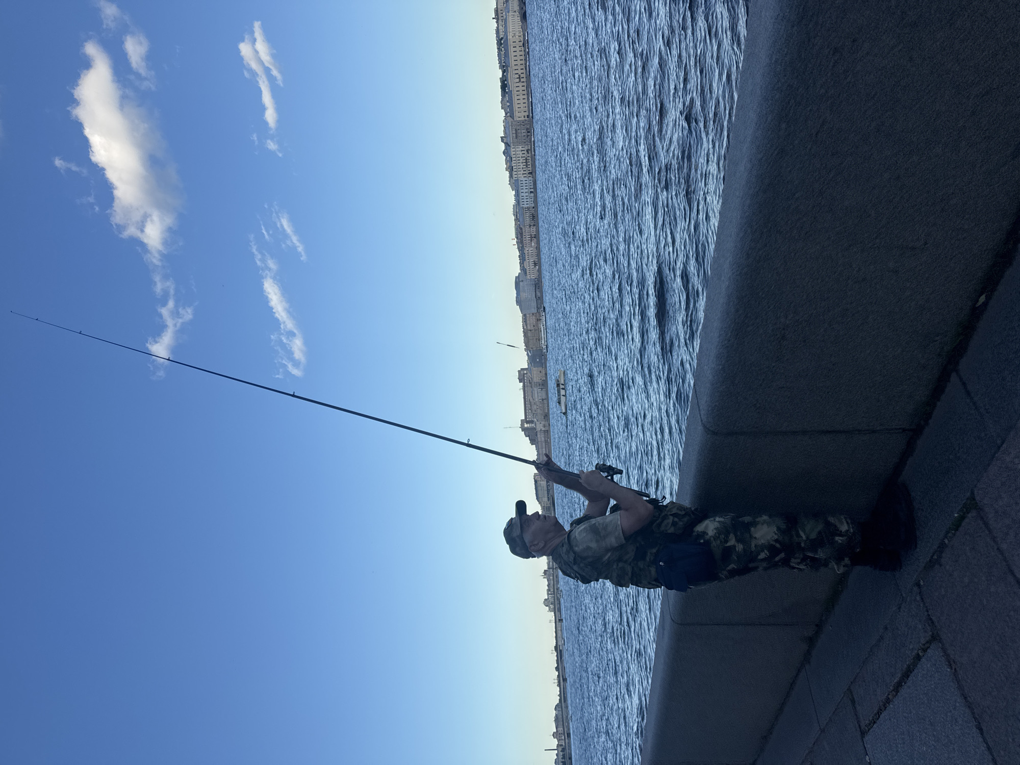 An elderly man stands on a stone embankment facing a wide river, fishing with a long rod under a clear blue sky with scattered clouds. He is wearing a camouflage vest over a green shirt, camouflage pants, a cap, and dark boots. A small dark bag is strapped to his waist. The river is choppy and a long fishing line extends upwards. Across the river, a city skyline with numerous buildings stretches along the opposite bank, with a few boats visible on the water. The embankment is paved with large stones. The scene appears to be a waterfront area in Saint Petersburg, Russia, likely on a sunny day.