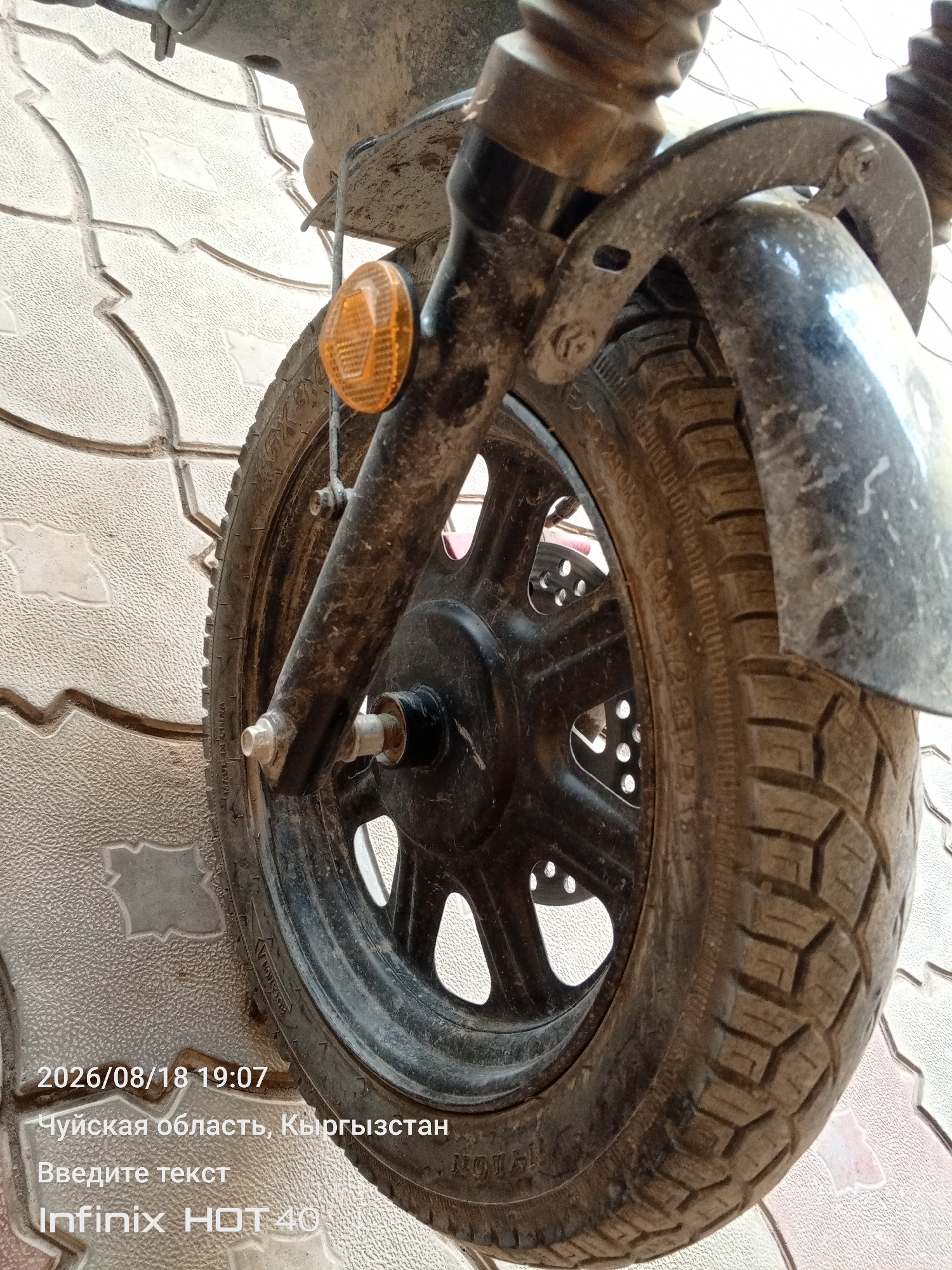 This is a close-up, low-angle, outdoor photograph of a dirty, black motorcycle wheel with a textured tire tread. The wheel is black and has several circular cutouts. The tire is dark gray and shows some wear. The motorcycle's front suspension and fender are visible, also covered in dust and dirt. An orange reflector is attached to the suspension. The background consists of a light-colored tiled surface with a diamond pattern. The photograph is dated 2026/08/18 19:07 and includes text overlays indicating "Chuy Oblast, Kyrgyzstan", "Enter text", and "Infinix HOT 40". The location of Alga, Kyrgyzstan is noted.
