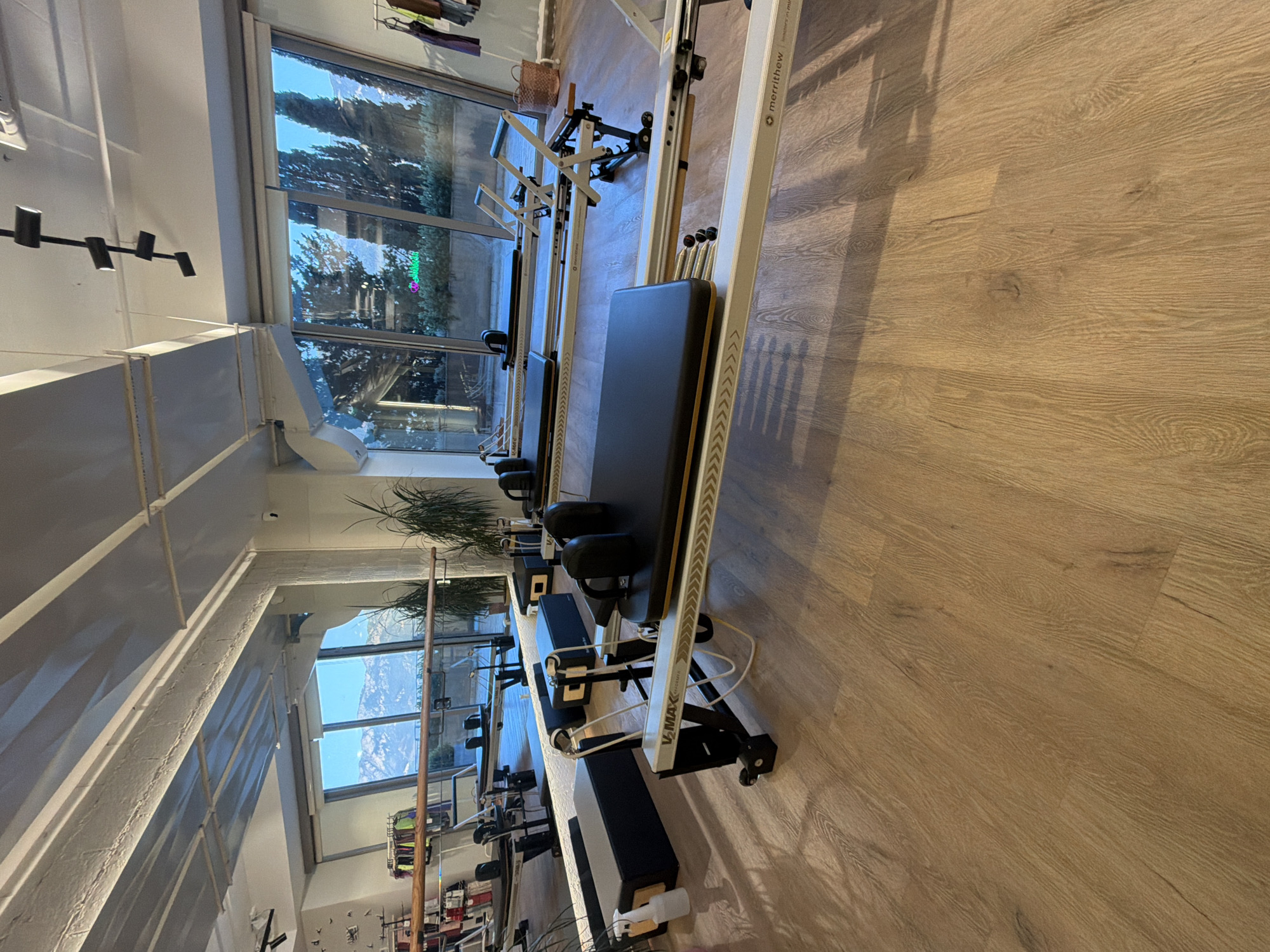 This is a medium-shot, eye-level, wide-angle photograph of a Pilates studio in Boreti, Montenegro, taken indoors during daylight. The studio has a polished wooden floor and large windows offering a view of snow-capped mountains and trees. Several Pilates reformers, which are large, specialized exercise machines, are arranged in the room. In the foreground, a white reformer with a black carriage and headrest is prominent, featuring the "V-MAX REFORMER" branding. To its right, another reformer is visible, with the "Merritthew" logo on its side. In the background, two more reformers are positioned facing the windows, and a tall plant is situated near the right window. The room is illuminated by natural light from the windows and artificial light from ceiling fixtures. There are no people visible in the scene.