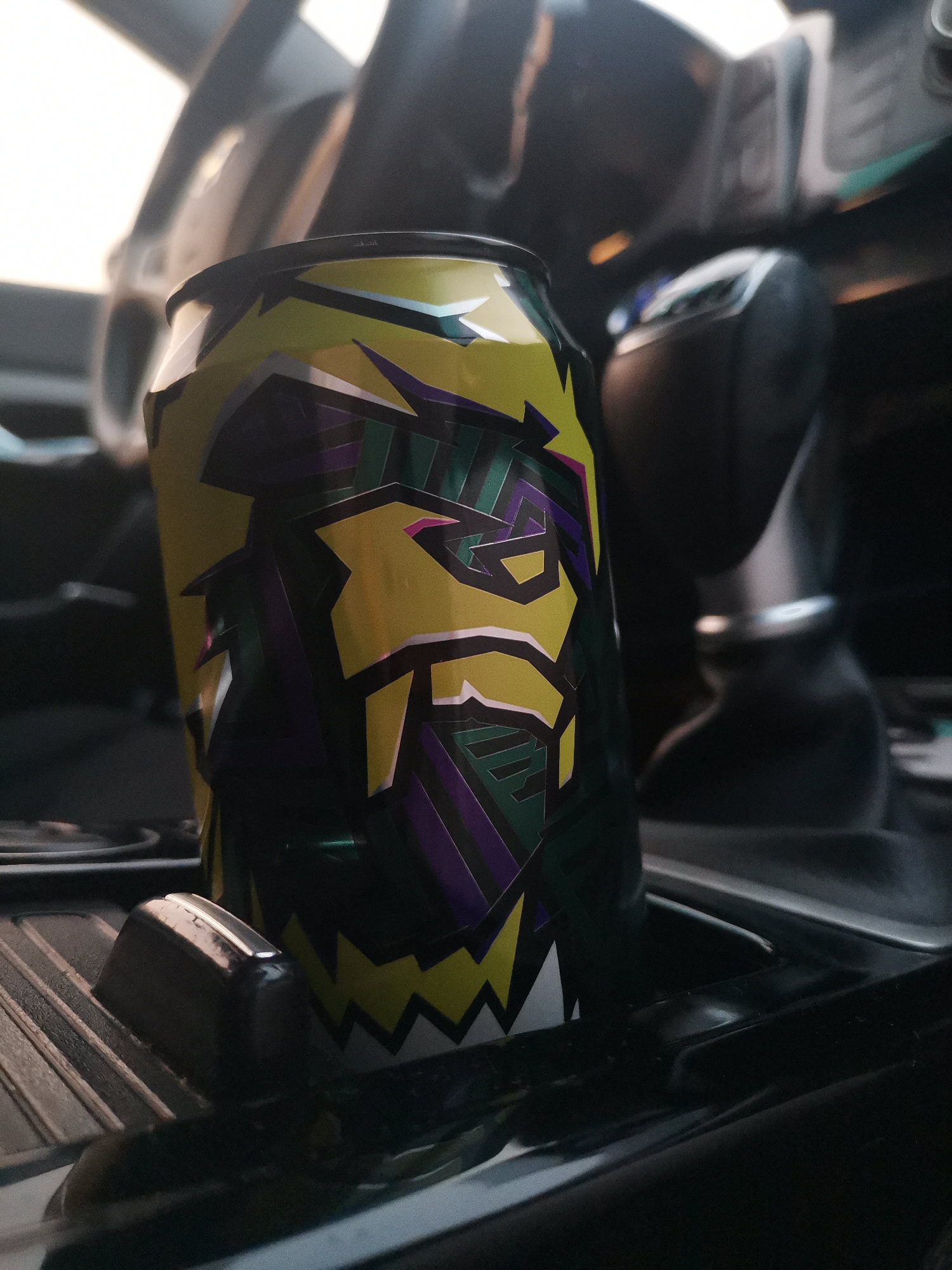 A can with a green, yellow, and purple abstract design is placed in a car's cup holder. The can is slightly tilted and the design appears to depict a stylized face or creature with sharp angles and geometric shapes. The background is blurred, showing parts of the car's interior, including what appears to be a gear shift and dashboard. The lighting suggests it is daytime. The location is not determinable from the image.
