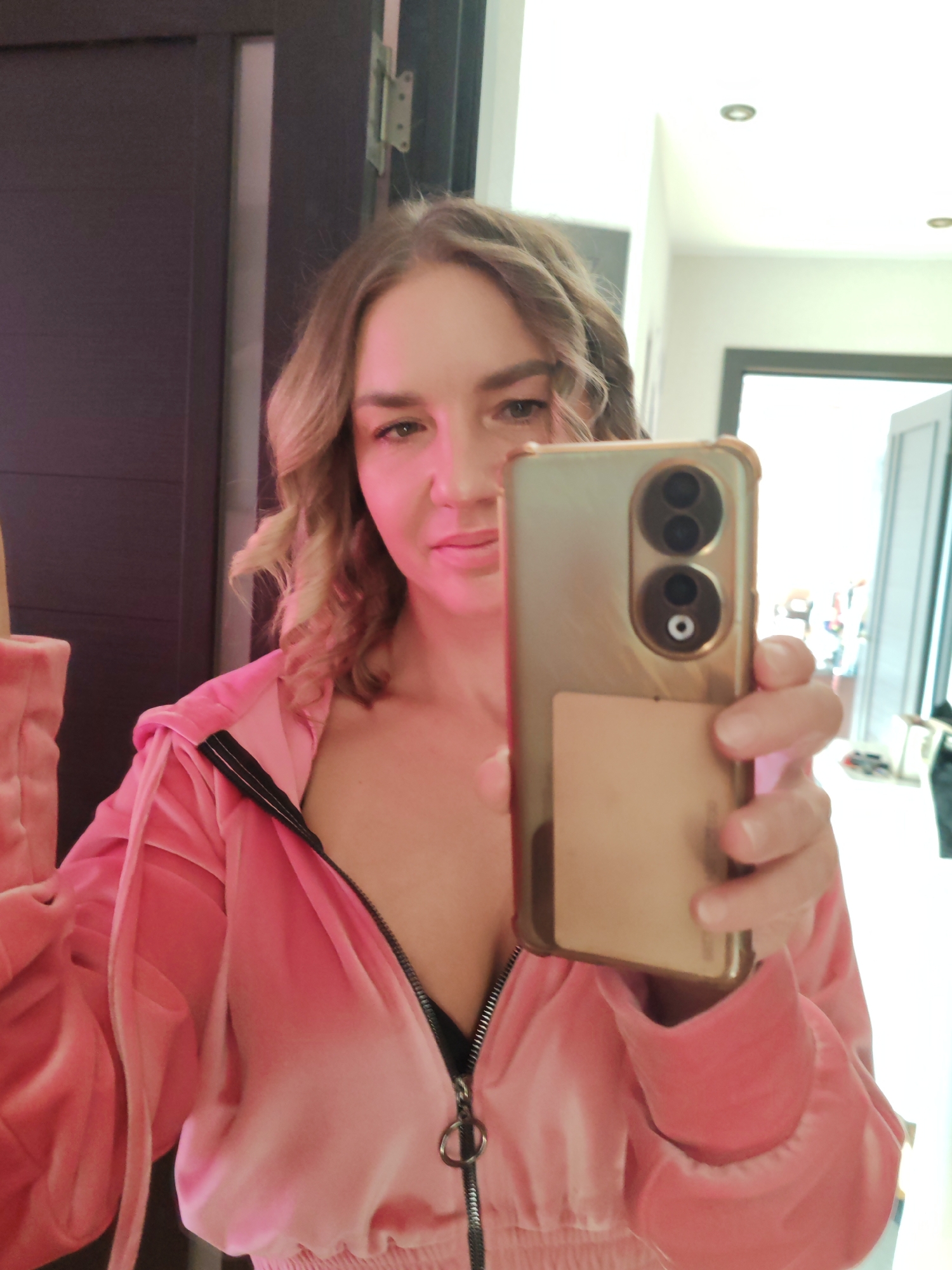A Caucasian woman with blonde, wavy hair is taking a selfie indoors. She is wearing a pink velvet zip-up jacket, partially unzipped to reveal a black top underneath. She holds a gold-colored smartphone up to the camera, obscuring part of her face.
The background features a dark-colored door to the left and an open doorway leading to a bright, naturally lit room on the right. The specific location in Petropavlovsk-Kamchatsky, Russia cannot be determined from this image.