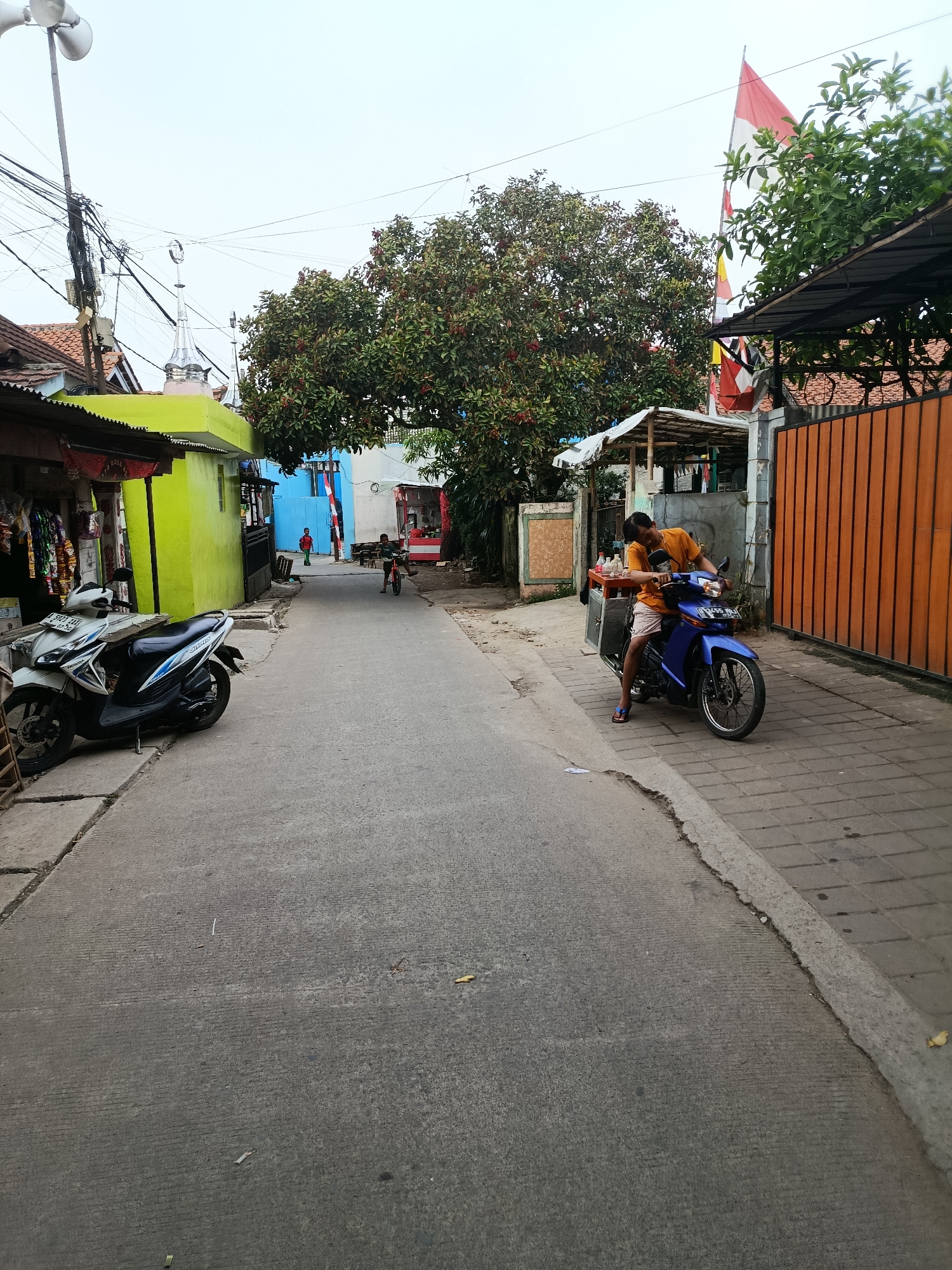 The image depicts a street scene in Balaraja, Indonesia. A man in an orange shirt and light-colored shorts sits on a blue motorcycle, leaning forward. The motorcycle has a food cart attached to its rear. The street is paved with asphalt, and there is a paved sidewalk on the right side of the image. Buildings with brightly colored facades, including a lime green one on the left and a blue one further down, line the street. A large tree with dense foliage stands in the middle ground. Several motorcycles are parked on the left side of the street. Two children are visible in the distance: one on a bicycle and another standing on the sidewalk. Indonesian flags are flying from poles on the right side of the image, near a tall orange wooden fence. The sky is overcast.