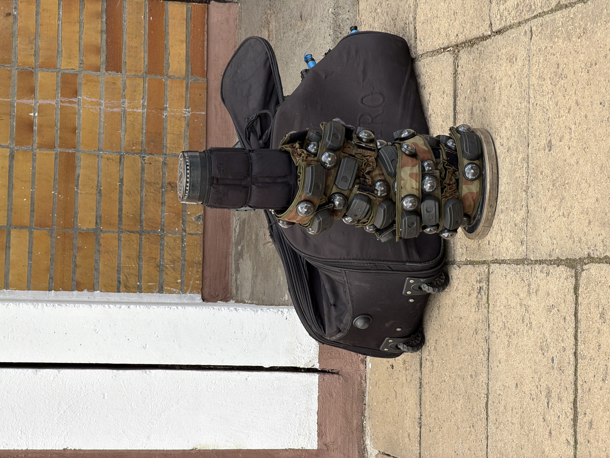 The image displays a large, cylindrical piece of equipment standing upright on a paved surface next to a brick wall. This primary object is predominantly black, with its mid-section wrapped in camouflage fabric featuring patterns of brown, green, and black. Numerous small, black, rectangular modules, each topped with a glossy, silver, hemispherical element (likely sensors or lights), are affixed around the camouflage section. The top of the cylindrical device is black with a textured or patterned ring.
Adjacent to this device lies an open, black, soft-sided equipment case, resembling a gig bag or gear bag. The word "PRO" is faintly visible in large lettering on its side. Several blue-tipped items, possibly parts of airsoft or paintball markers, are seen protruding from the case. The case itself features wheels and reinforced corners, indicative of a transport bag.
The setting appears to be outdoors on a clear day, with natural light illuminating the scene. The ground is a light-colored paved surface, made of rectangular concrete blocks or pavers. To the left of the main objects, there is a brown-painted border and a distinct white painted structure, possibly a ledge or step, with a dark horizontal line separating two white sections. Further to the left and behind, a wall constructed from light orange and brown rectangular bricks with white mortar lines is visible. There are no people in the image. The collection of items suggests equipment used for a tactical or recreational game, such as laser tag or paintball.