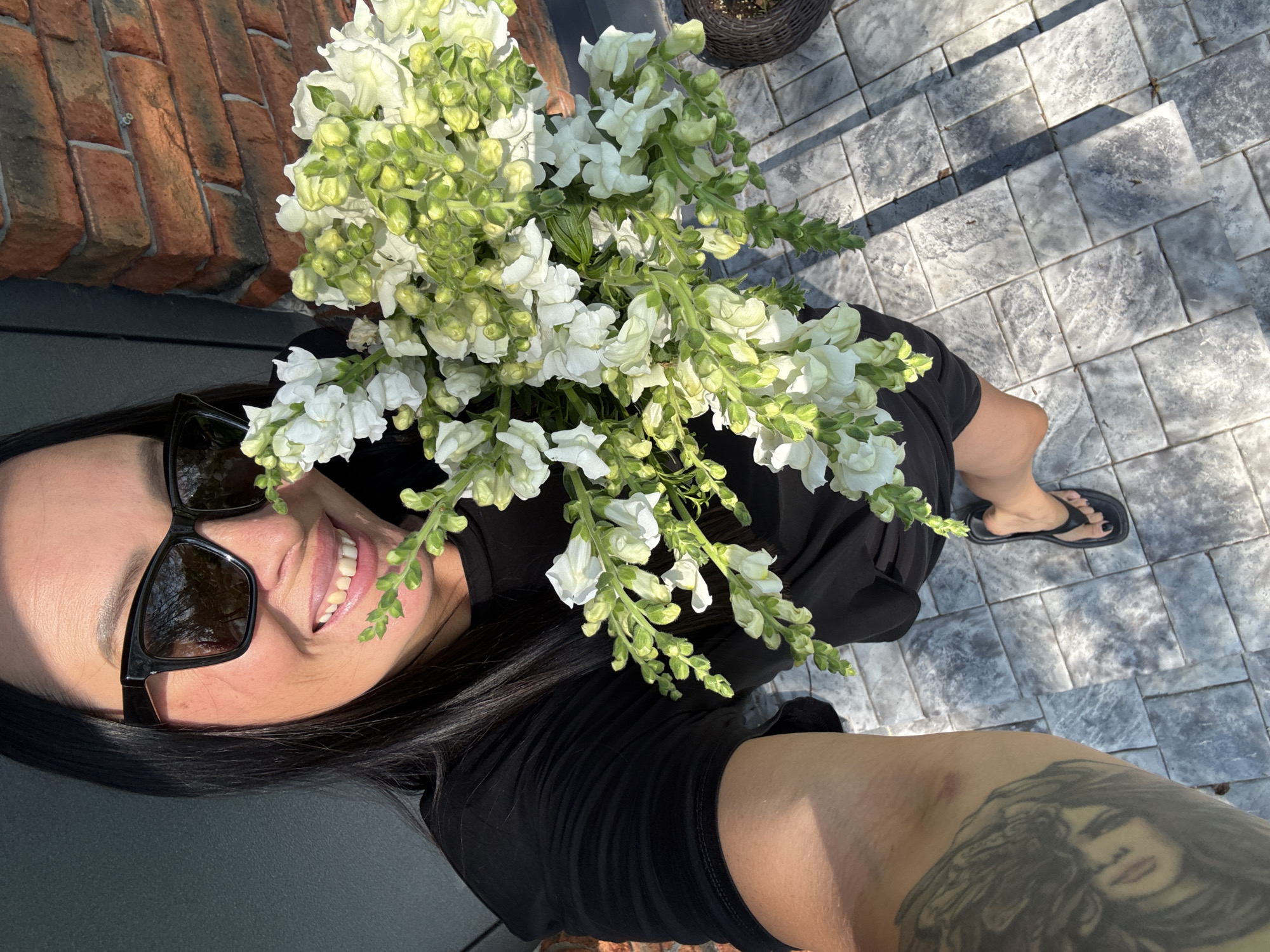 A woman with long, dark hair and black sunglasses is taking a selfie. She is smiling and holding a bouquet of white snapdragon flowers. She is wearing a black t-shirt and black flip-flops. She has a large tattoo on her left arm. The background shows a brick wall and a gray tiled patio. The image does not contain any information about the city of Chaikovsky, Russia.