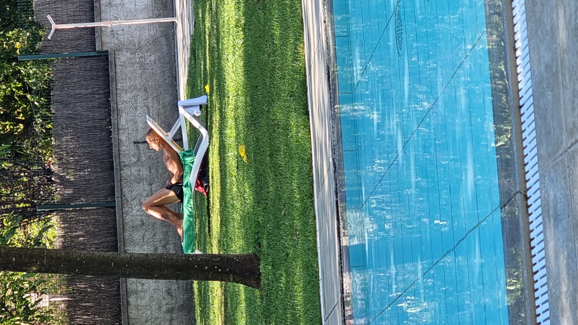 A man is sunbathing on a white lounge chair on a green lawn. He is wearing black swim trunks and appears to be naked from the waist up. The lounge chair is partially covered by a green towel and a red bag. In the background, there is a grey stone wall and a wooden fence. A tall, thin, white object, possibly a light fixture or a flag pole, stands to the right of the lounge chair. A tree trunk is visible on the left side of the frame. The foreground shows the edge of a blue swimming pool with reflections of trees on its surface. The overall scene suggests an outdoor recreational area, likely a backyard or a poolside setting. The location is stated to be Sant Joan Les Fonts, Spain.