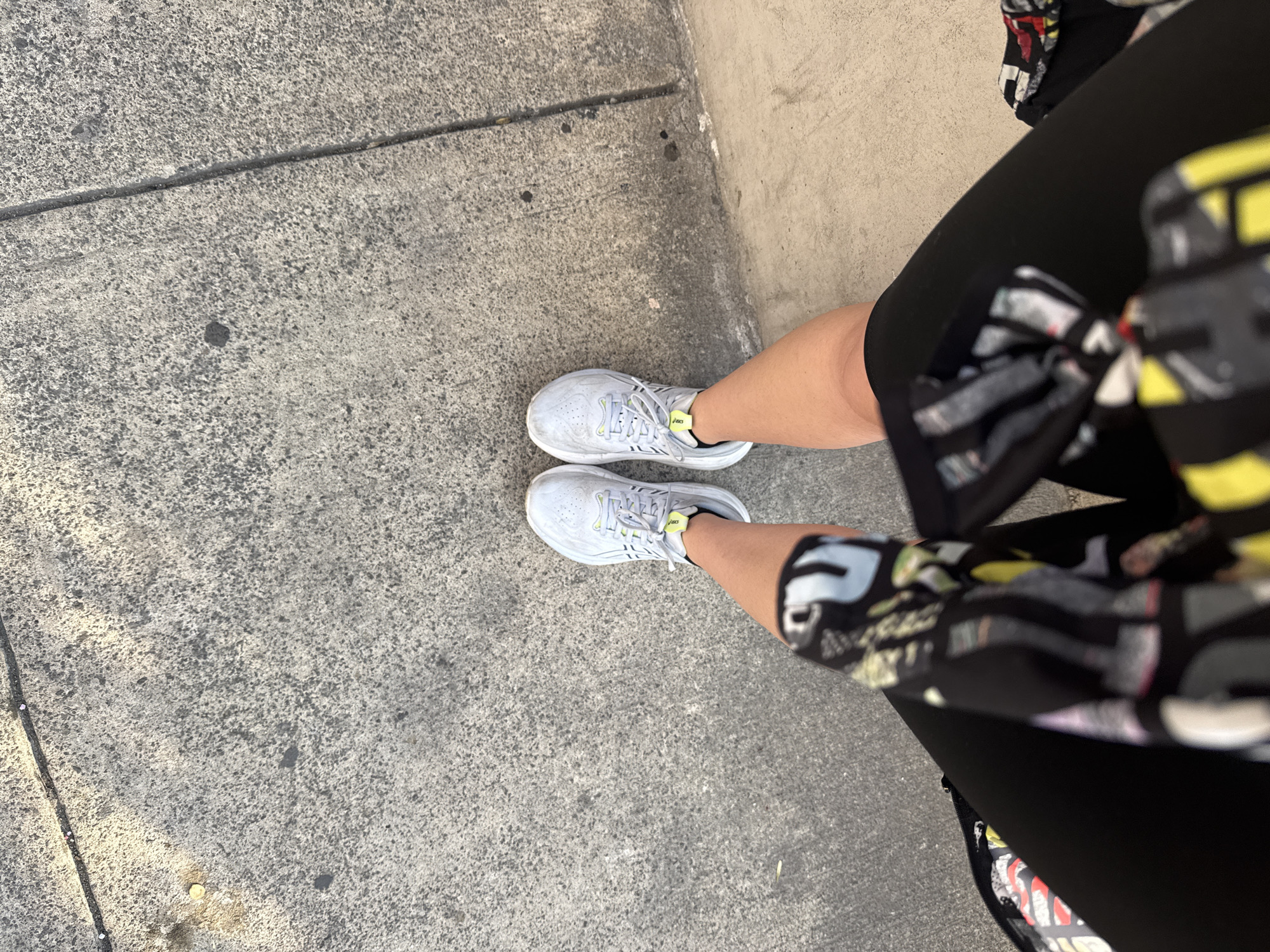 A high-angle shot shows a person standing on a concrete sidewalk. The person is wearing black leggings, a black, gray, and yellow patterned top, and gray ASICS running shoes with yellow accents. The sidewalk is cracked and stained in places. The location is most likely in Mexico City, Mexico, given the description provided.